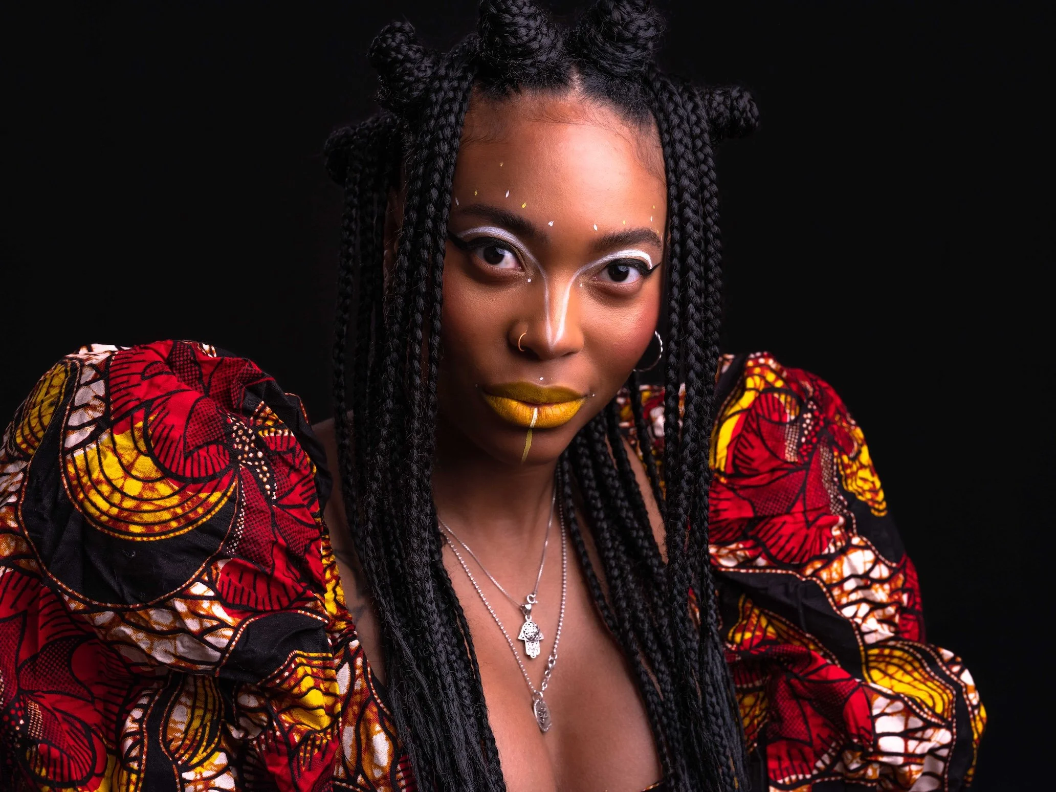 Professional Portrait Photography by Minneapolis Photographer Jay Cupcake. A woman with braided hair and gold makeup, wearing colorful African-patterned clothing and multiple silver necklaces, posing against a black background.