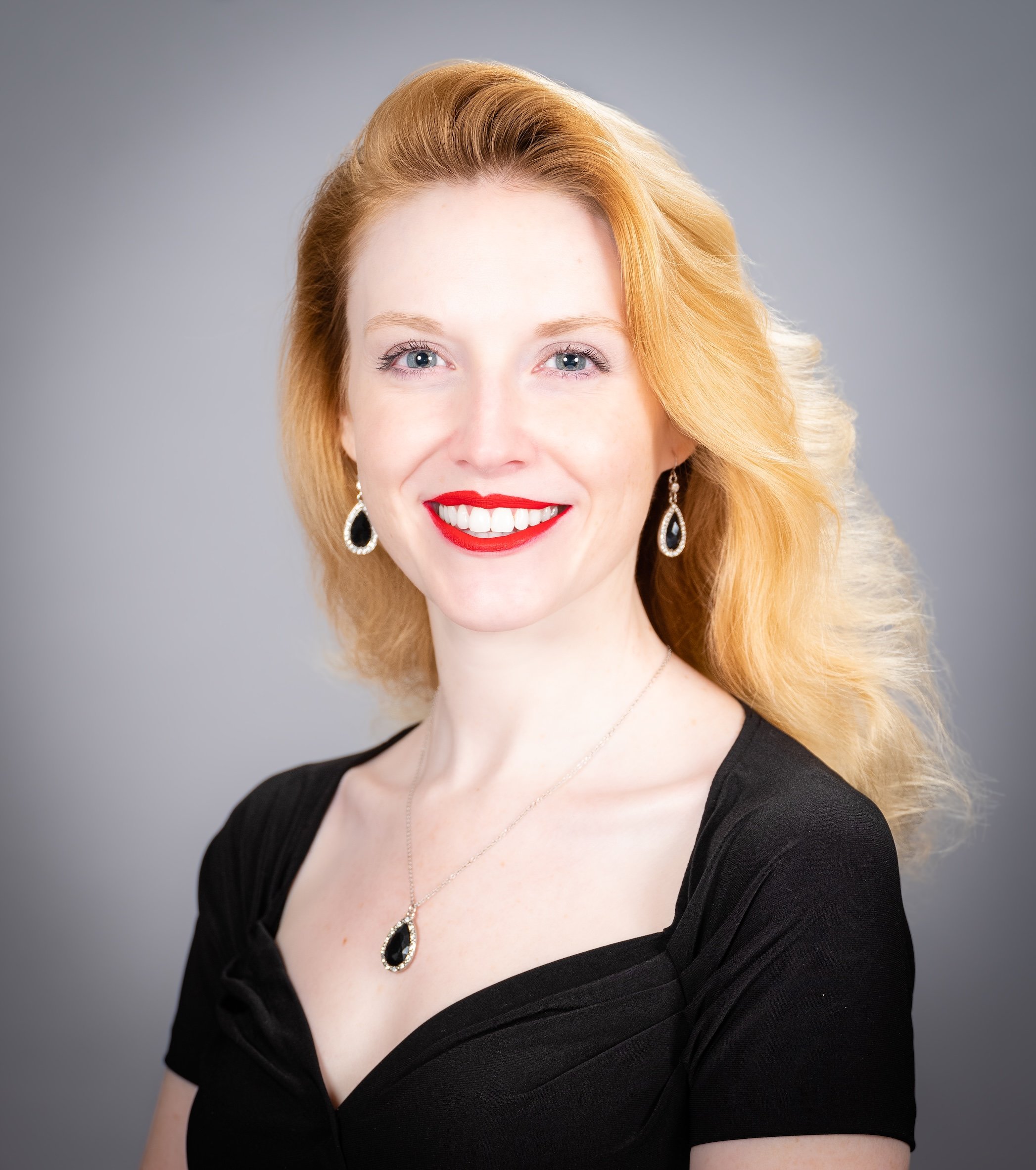 Professional Corporate Headshot by Saint Paul Photographer Jay Cupcake. Portrait of a woman with red hair, blue eyes, red lipstick, wearing earrings and a necklace with black and silver stones, dressed in a black top against a gray background.