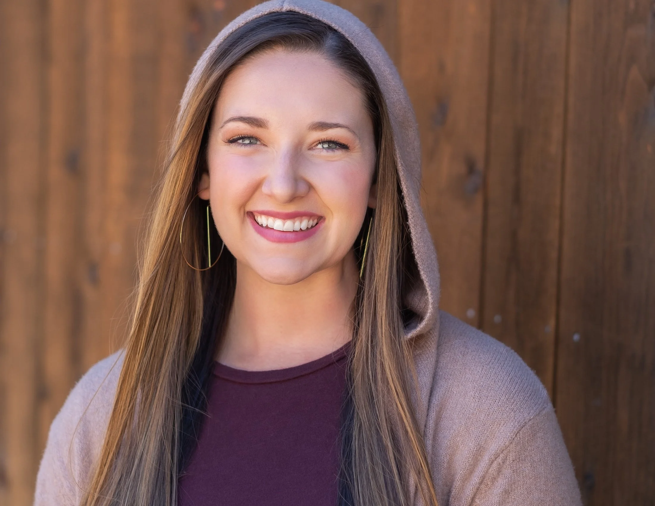 Professional Actor Headshot by Twin Cities Photographer Jay Cupcake. A young woman smiling with long blonde hair, blue eyes, wearing hoop earrings, a purple top, and a hoodie, standing in front of a wooden fence.