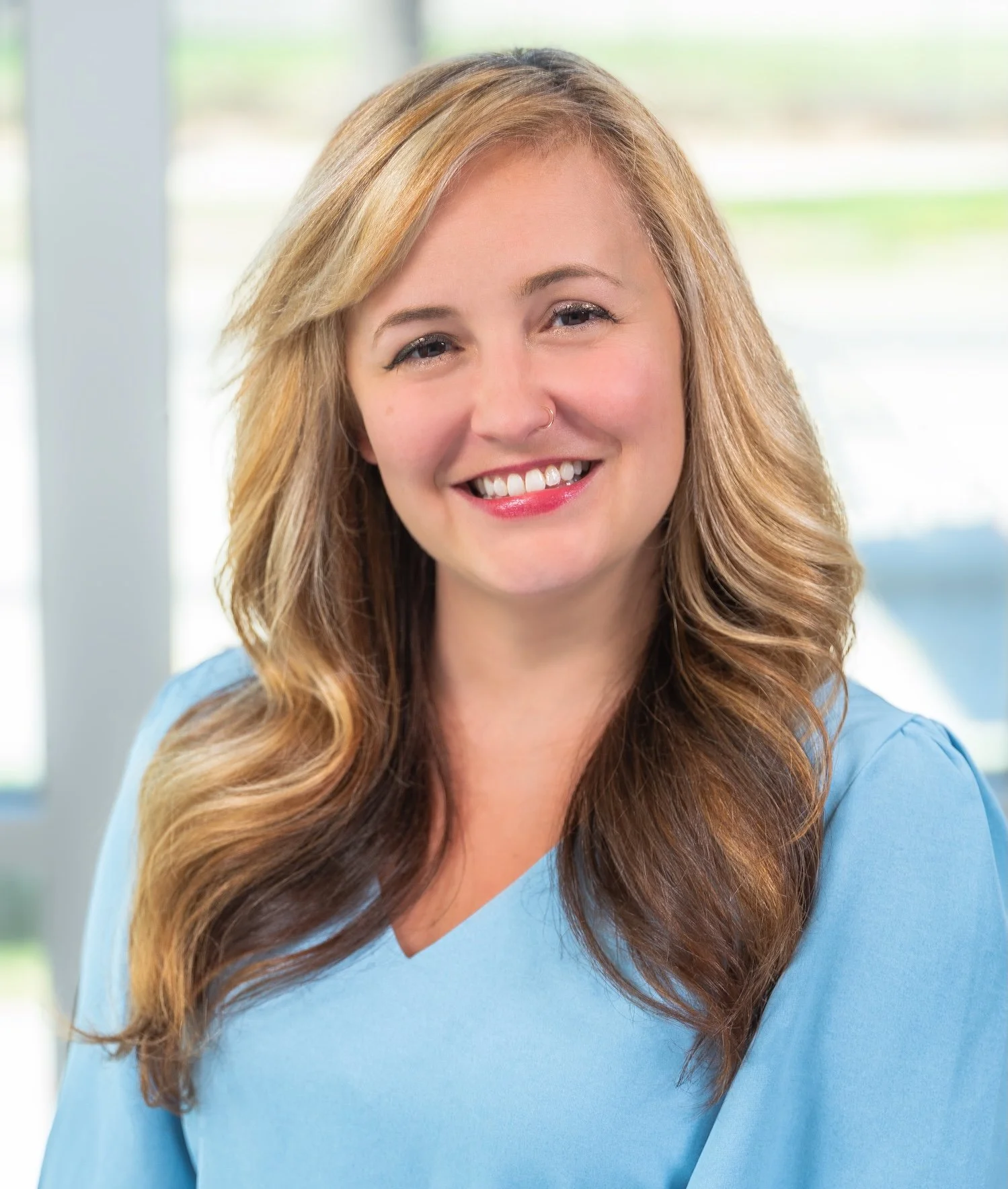 Professional Corporate Headshot by Minneapolis Photographer Jay Cupcake. A smiling woman with long, wavy blonde hair, wearing a light blue top, standing in front of a bright window.