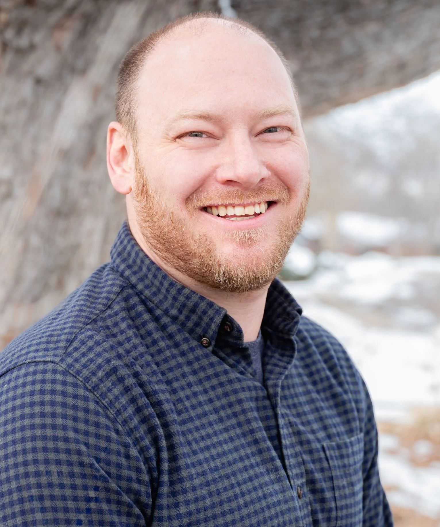 Professional Corporate Headshot by Twin Cities Photographer Jay Cupcake. A smiling man with a beard and light skin, wearing a dark blue checkered shirt, outdoors in a winter setting with snow and a large tree in the background.