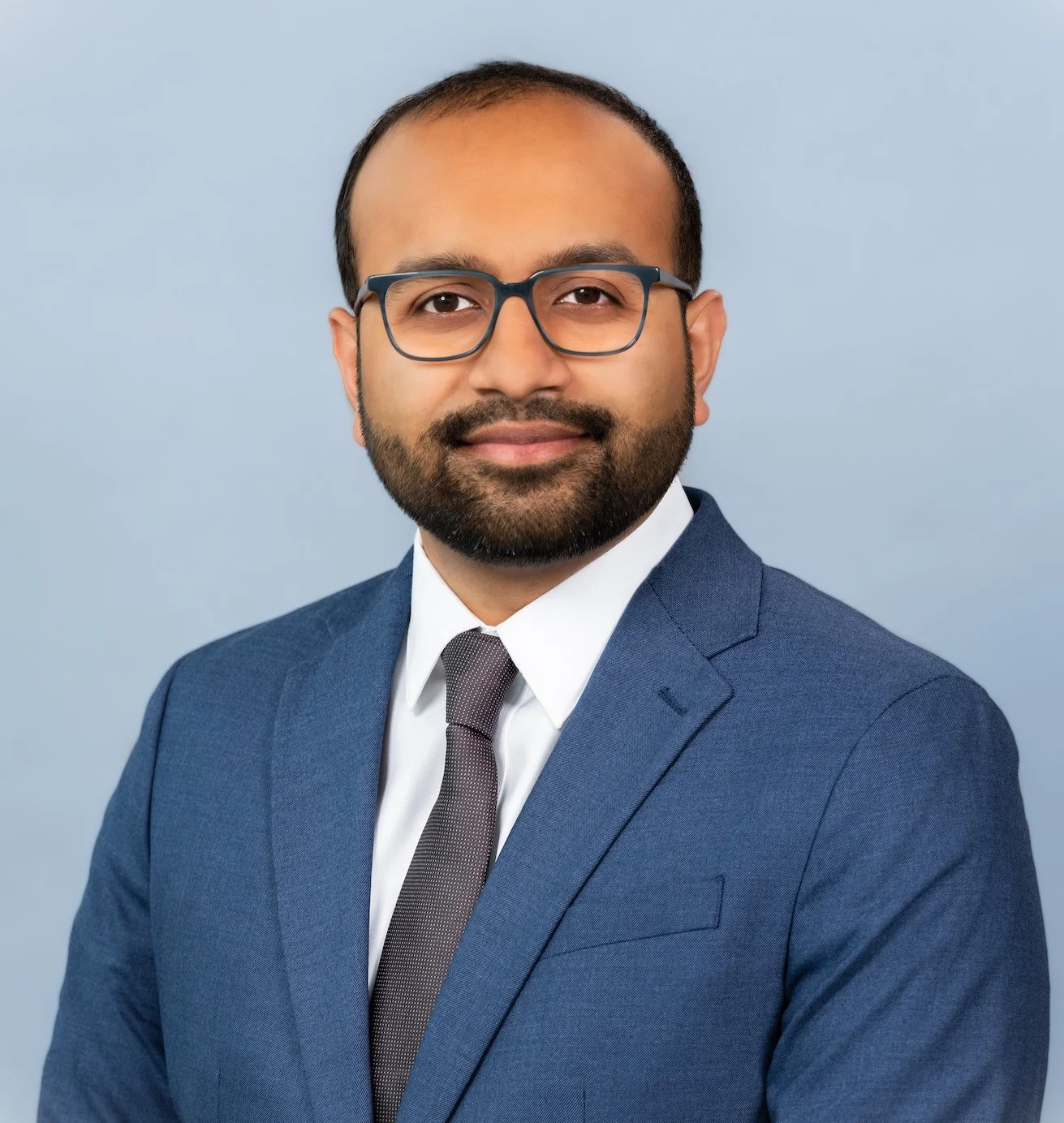 Professional Corporate Headshot by Saint Paul Photographer Jay Cupcake. Professional headshot of a man with glasses, a beard, wearing a blue suit and tie, against a light blue background.