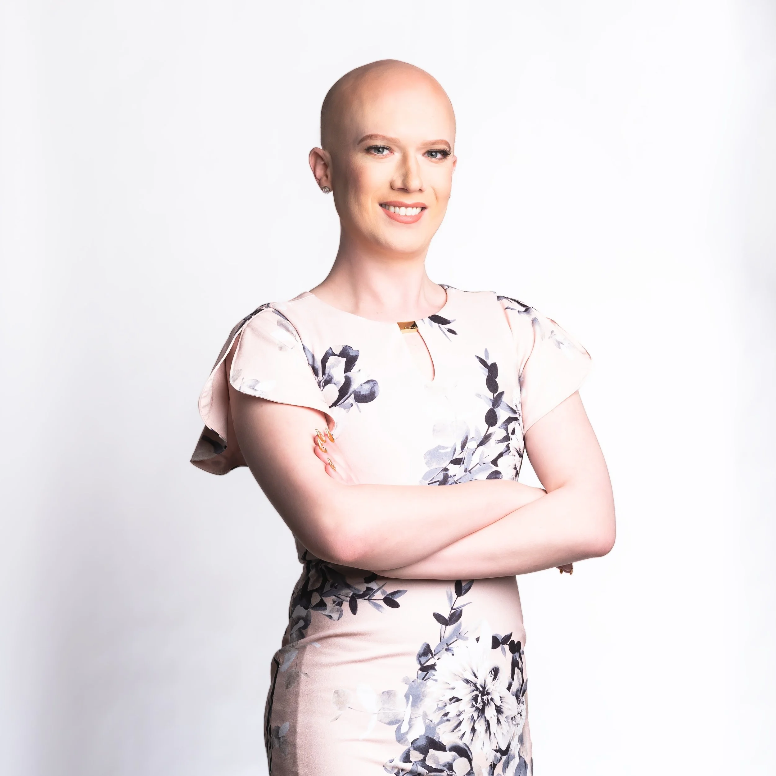 Professional Corporate Headshot by Twin Cities Photographer Jay Cupcake. A woman with a shaved head smiling and crossing her arms, wearing a floral dress against a plain white background.