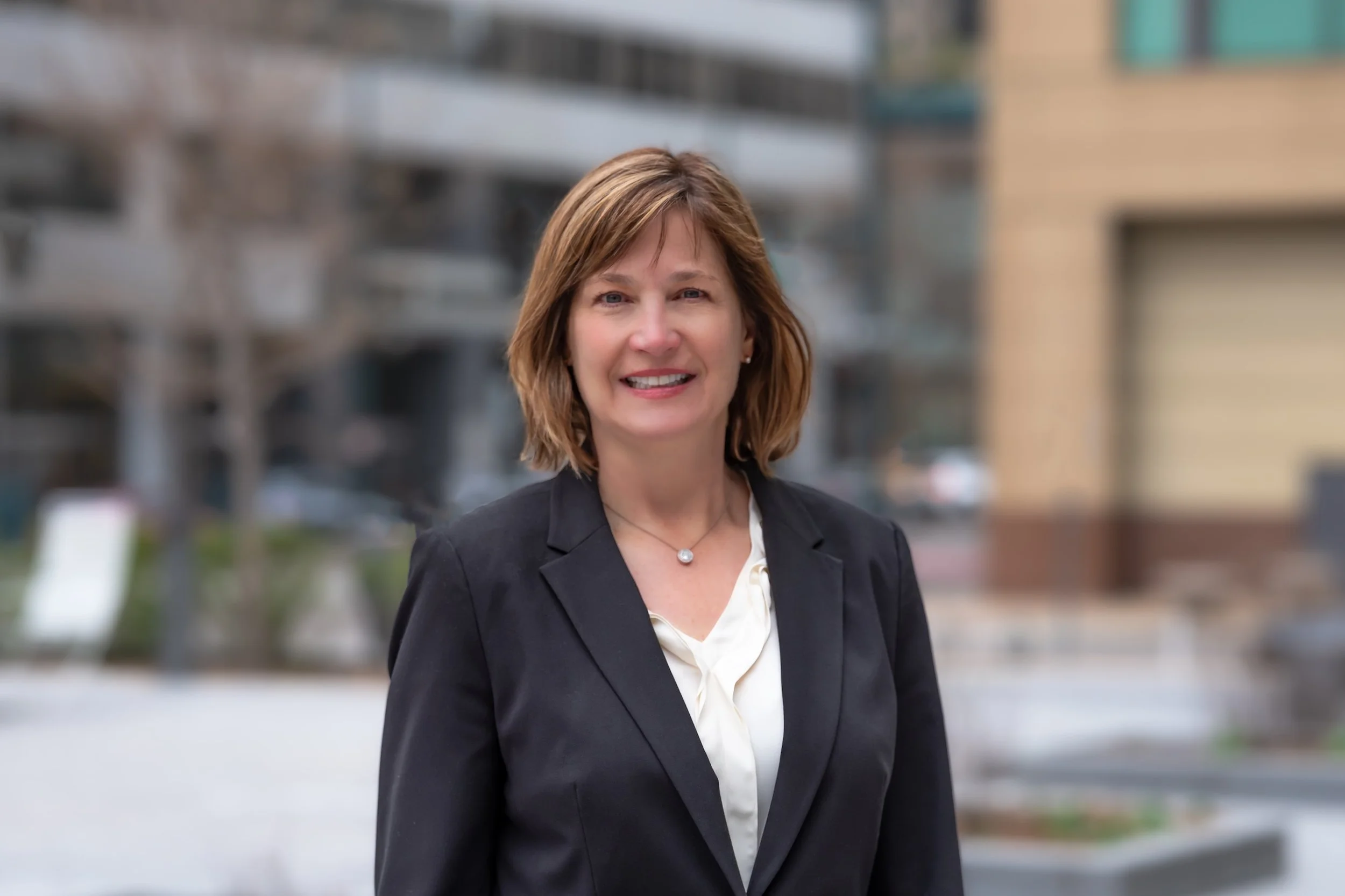 Professional Corporate Headshot by Saint Paul Photographer Jay Cupcake. Portrait of a smiling woman with shoulder-length brown hair, wearing a black blazer and a white blouse, standing outdoors in an urban area.