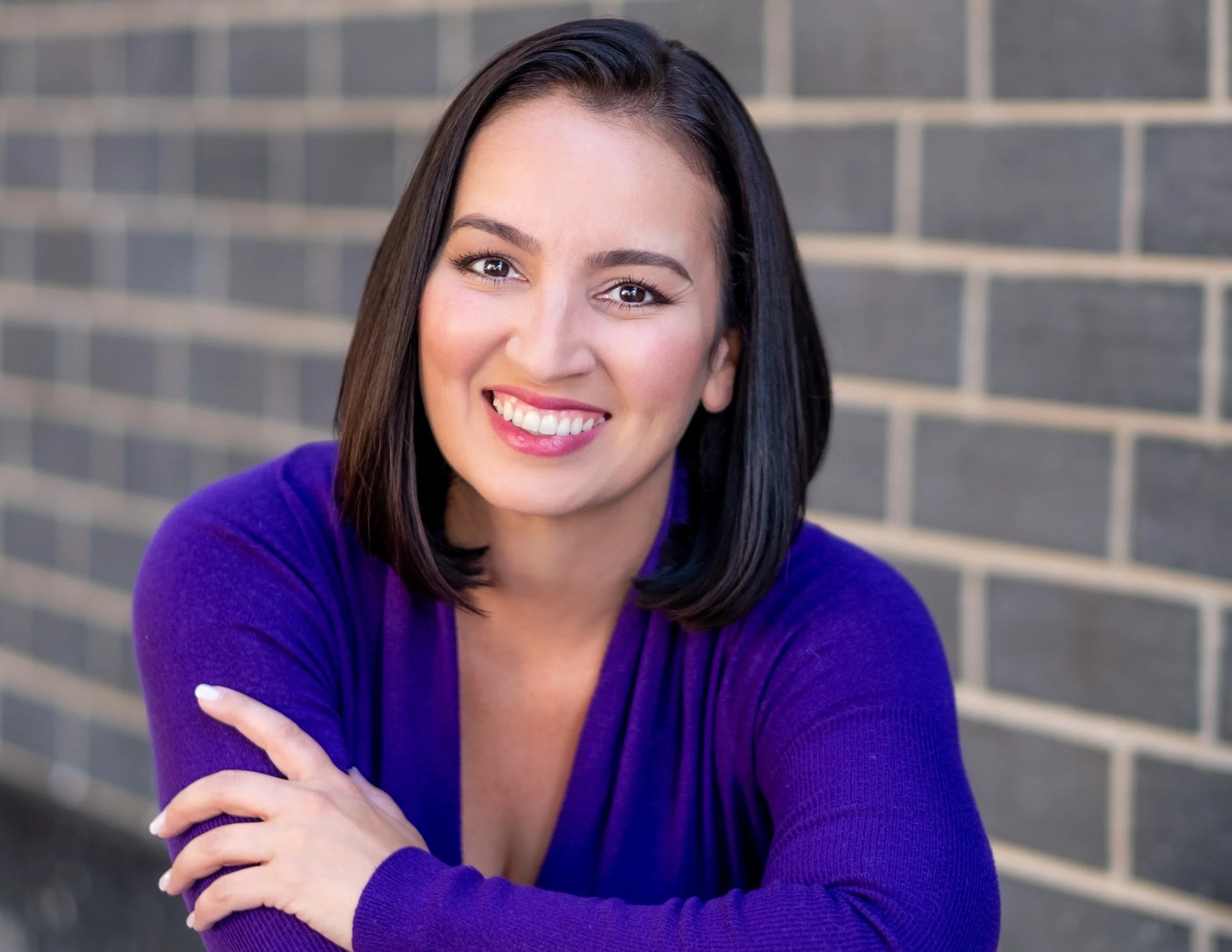 Professional Actor Headshot by Twin Cities Photographer Jay Cupcake. A young woman with shoulder-length dark hair smiling at the camera, wearing a purple sweater, with a brick wall background.