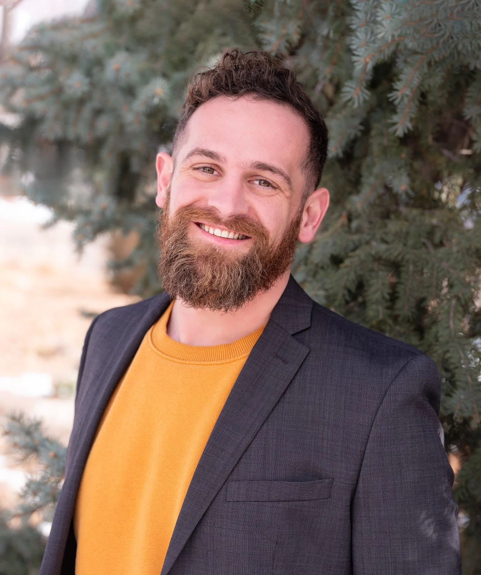 Professional Corporate Headshot by Minneapolis Photographer Jay Cupcake. A smiling man with a beard and short curly hair, wearing a black blazer and a mustard yellow sweater, standing outdoors near pine trees.