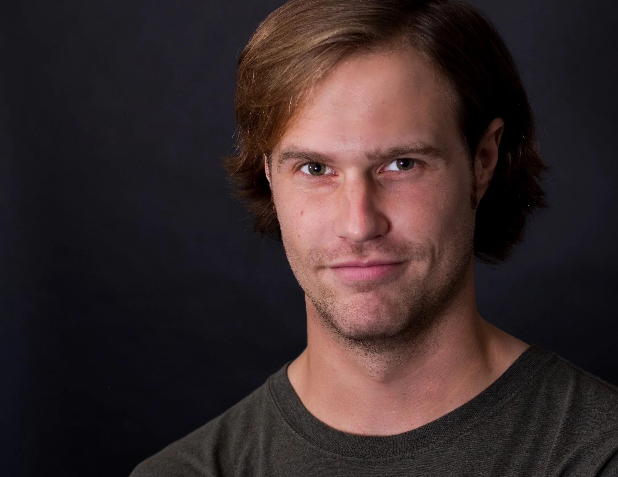 Professional Actor Headshot by Saint Paul Photographer Jay Cupcake. Close-up portrait of a young man with light brown hair, blue eyes, and a slight smile, wearing a dark gray t-shirt against a dark background.