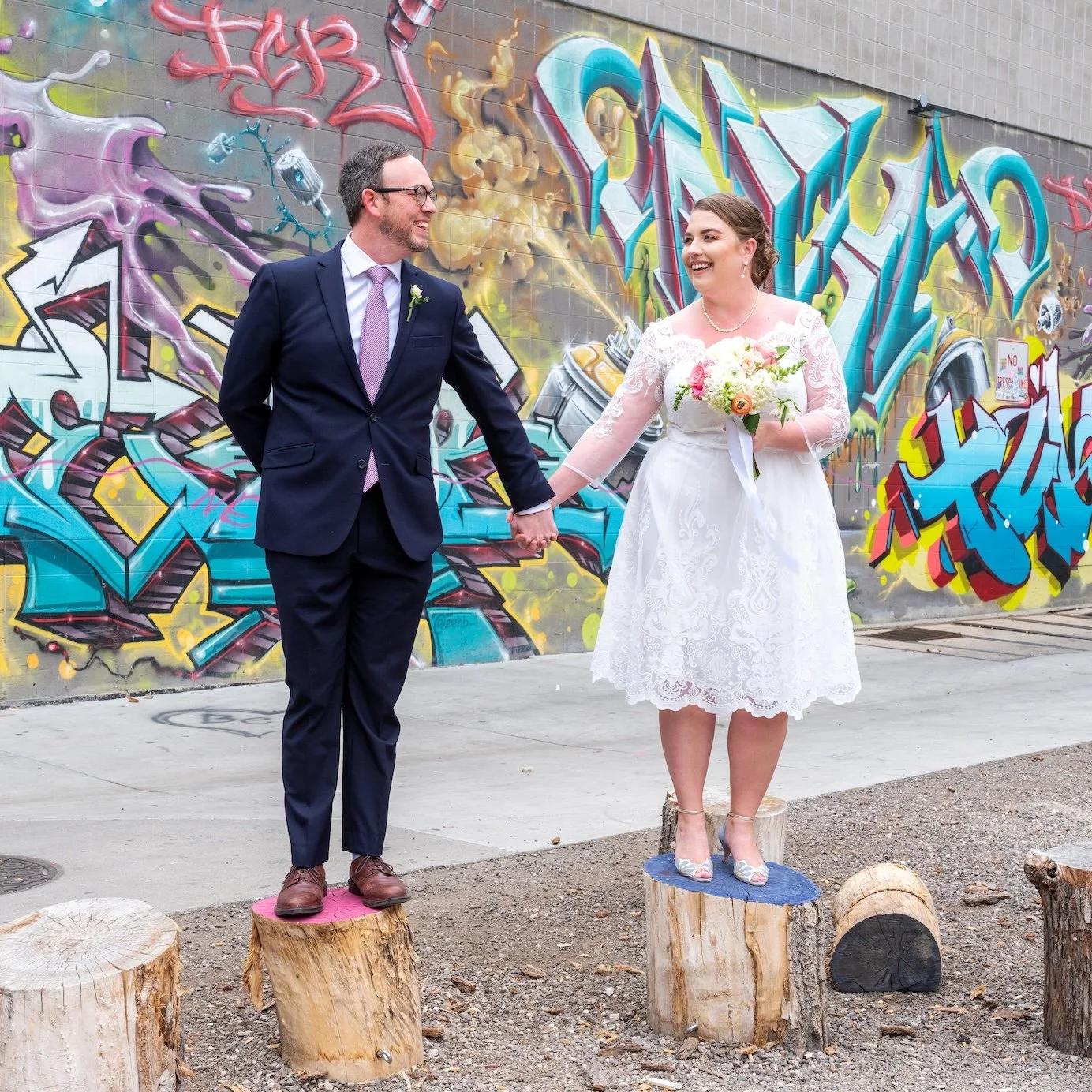 Professional Event Photography by Minneapolis Photographer Jay Cupcake. A bride and groom holding hands and smiling at each other during their wedding, standing on tree stumps in front of a colorful graffiti wall.