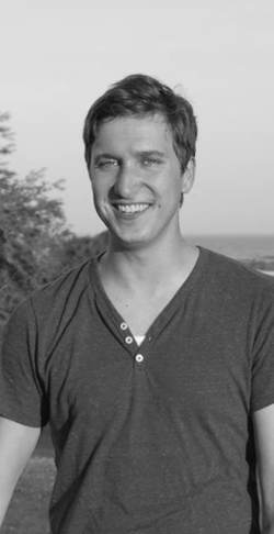Black and white photo of a smiling young man with short hair and a dark t-shirt, outdoors with trees and open sky in the background.