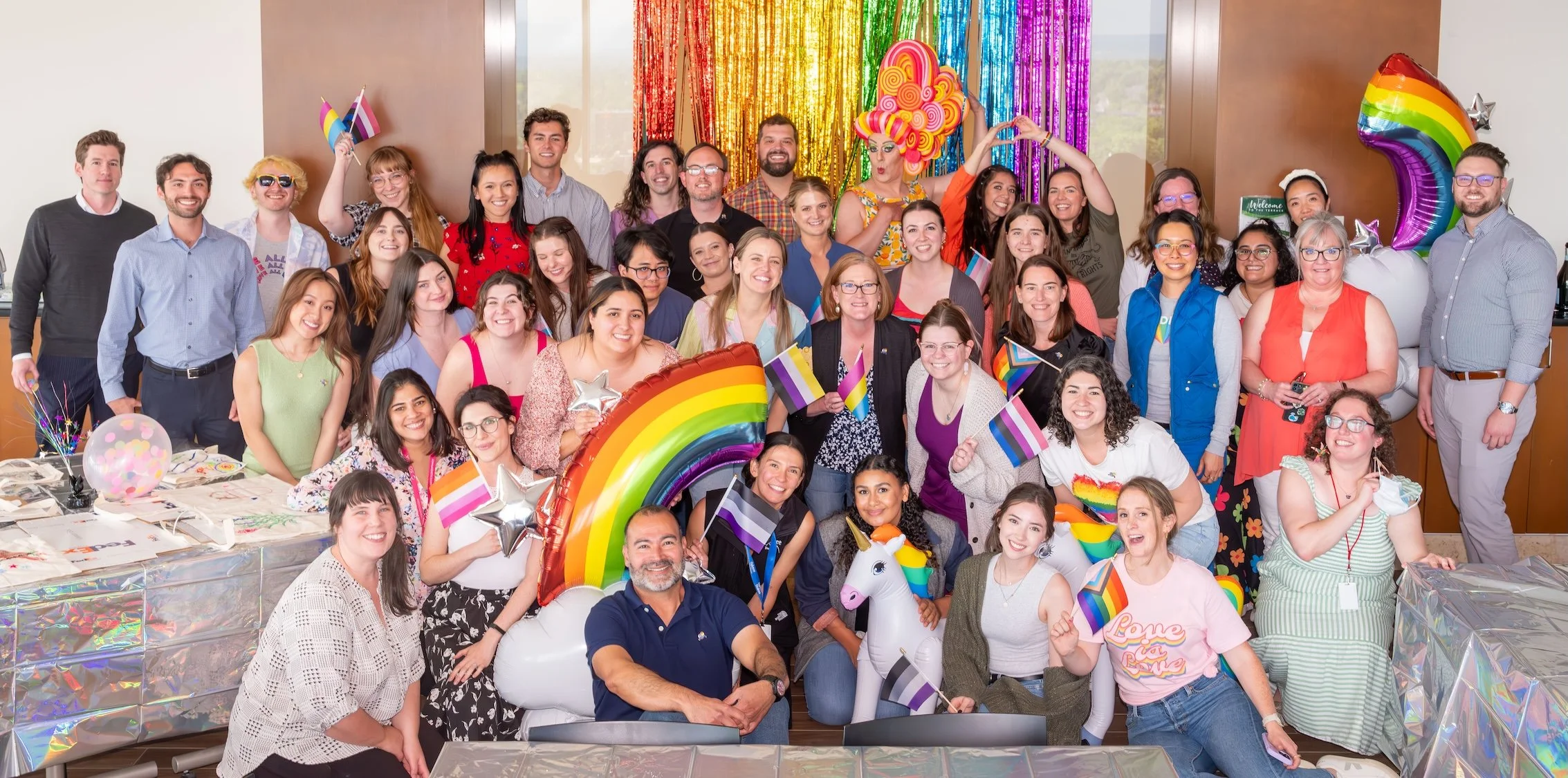 Professional Event Photography by Minneapolis Photographer Jay Cupcake. A diverse group of people gathered indoors celebrating with colorful rainbow-themed decorations, balloons, banners, and flags, including a rainbow balloon arch, rainbow unicorn, 