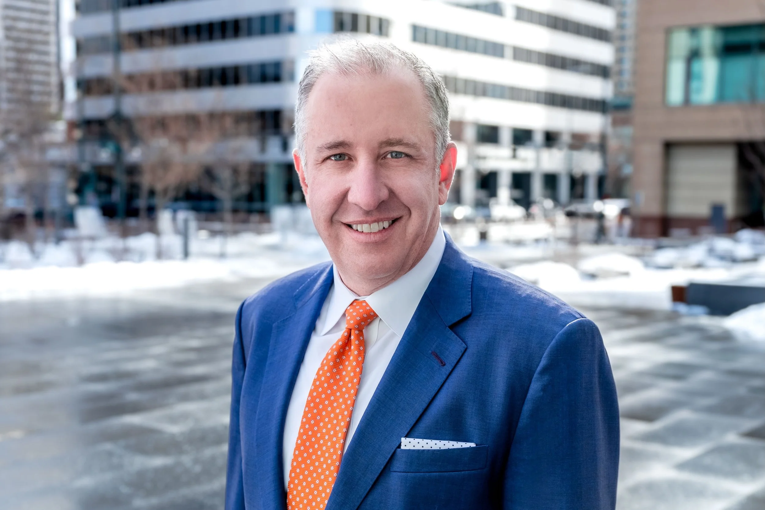Professional Corporate Headshot by Minneapolis Photographer Jay Cupcake. A smiling man in a blue suit and orange tie standing outside in a snowy urban setting with modern buildings in the background.