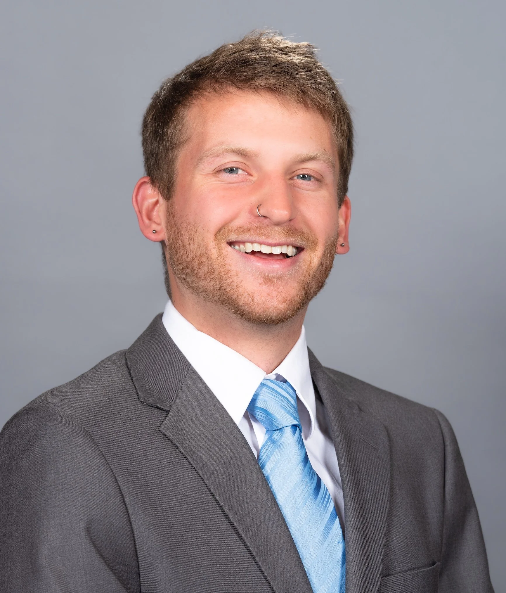 Professional Corporate Headshot by Twin Cities Photographer Jay Cupcake. Headshot of a smiling man with a beard, light brown hair, and blue eyes, wearing a gray suit, white shirt, and blue tie, against a gray background.
