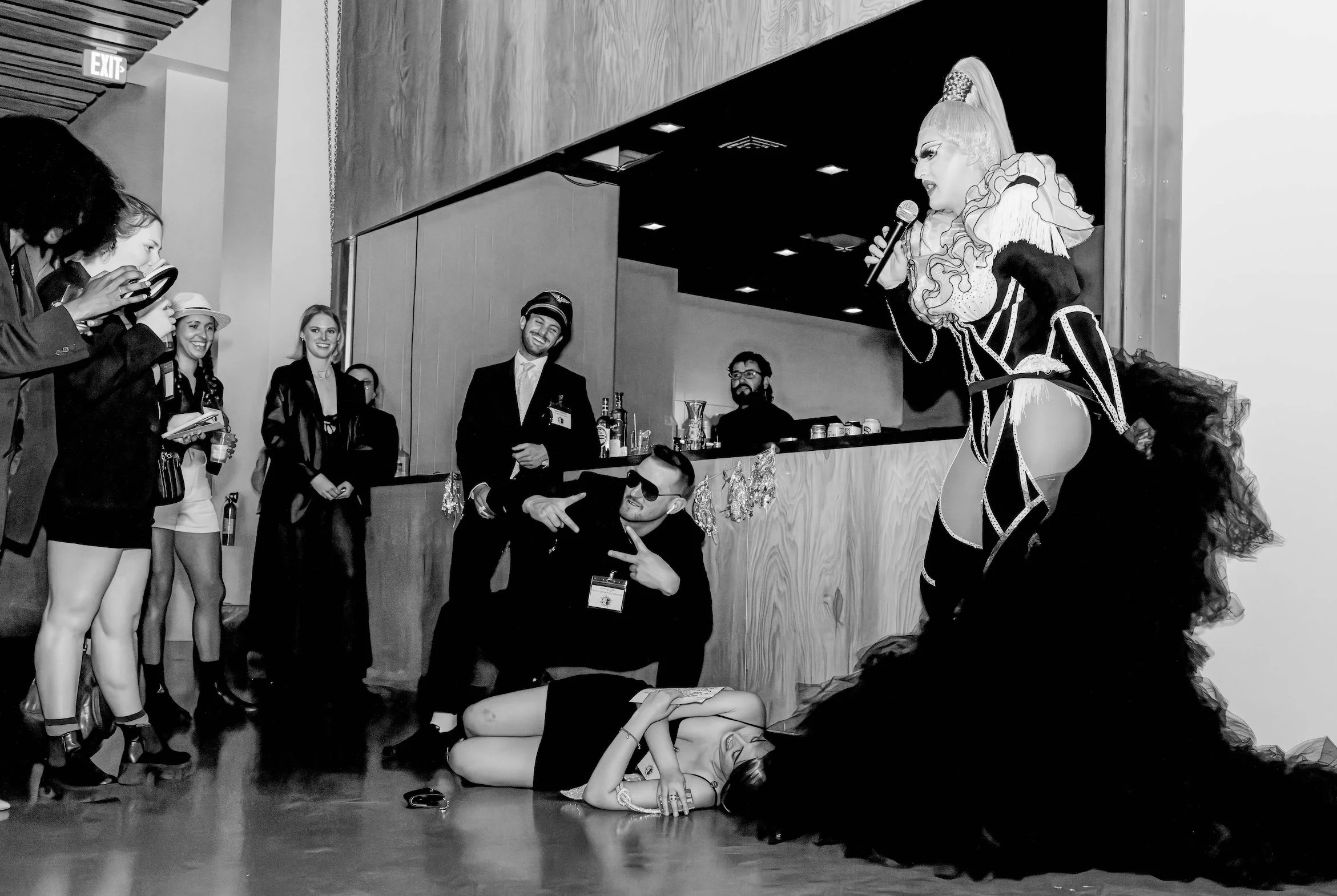 Professional Event Photography by Minneapolis Photographer Jay Cupcake. A performer dressed as a clown with a high ponytail and ruffled outfit singing into a microphone on a stage. A woman in a black dress is lying on the floor, covering her face. A 