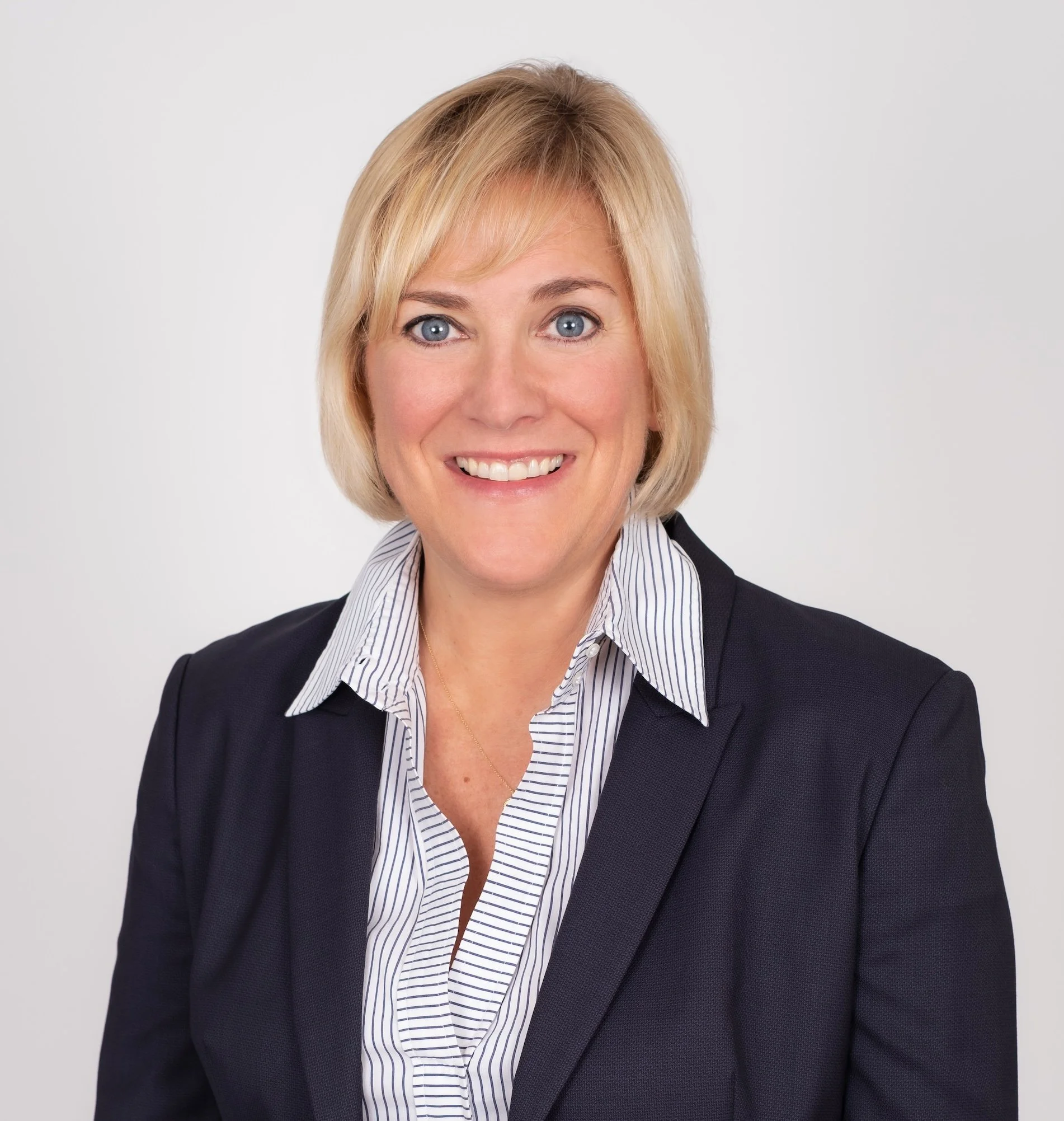 Professional Corporate Headshot by Minneapolis Photographer Jay Cupcake. A woman with blonde hair, blue eyes, and a friendly smile, dressed in a dark blazer and striped collared shirt, posing against a plain white background.