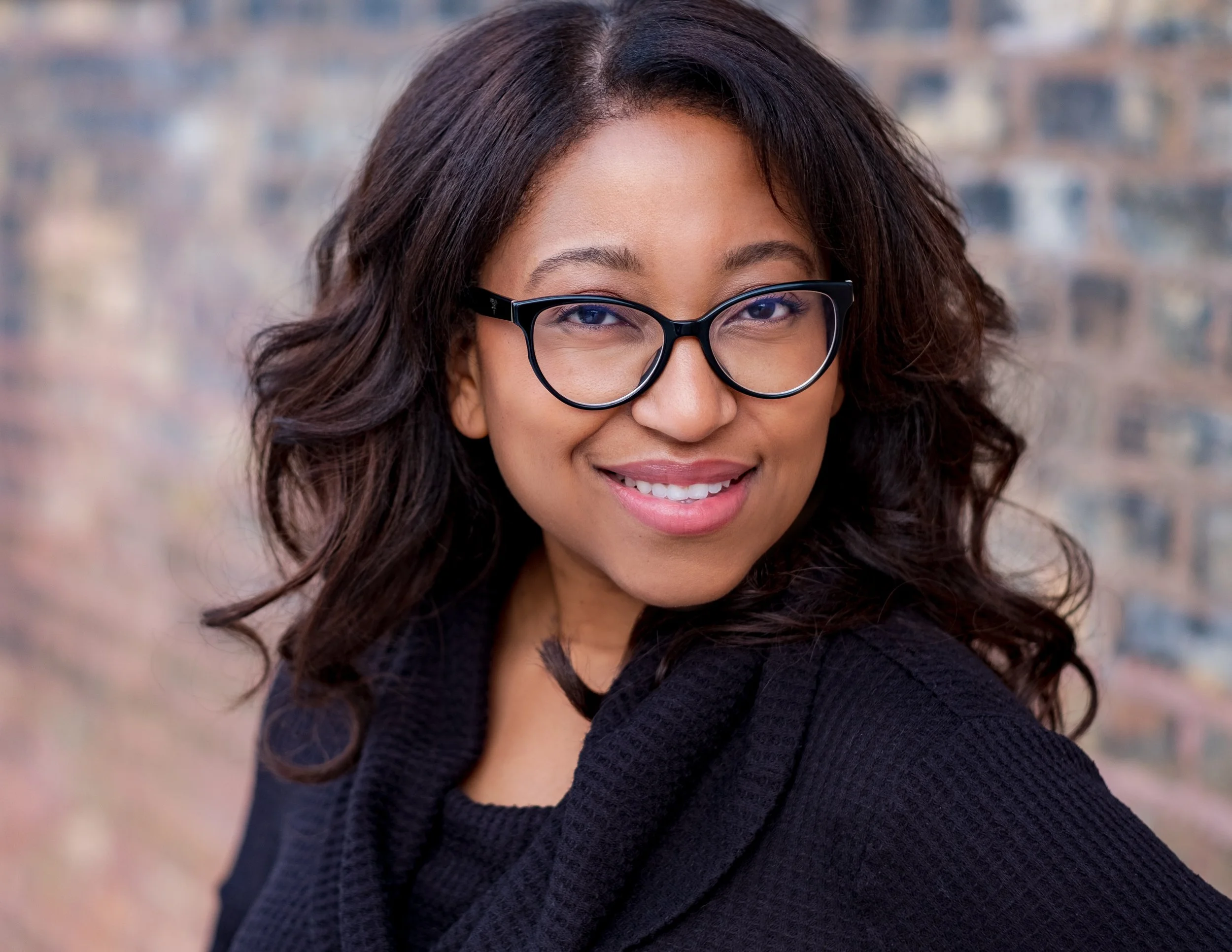 Professional headshot by St. Paul photographer Jay Cupcake. A smiling woman with glasses and wavy dark hair, wearing a black sweater, standing outdoors with a blurred cityscape background.