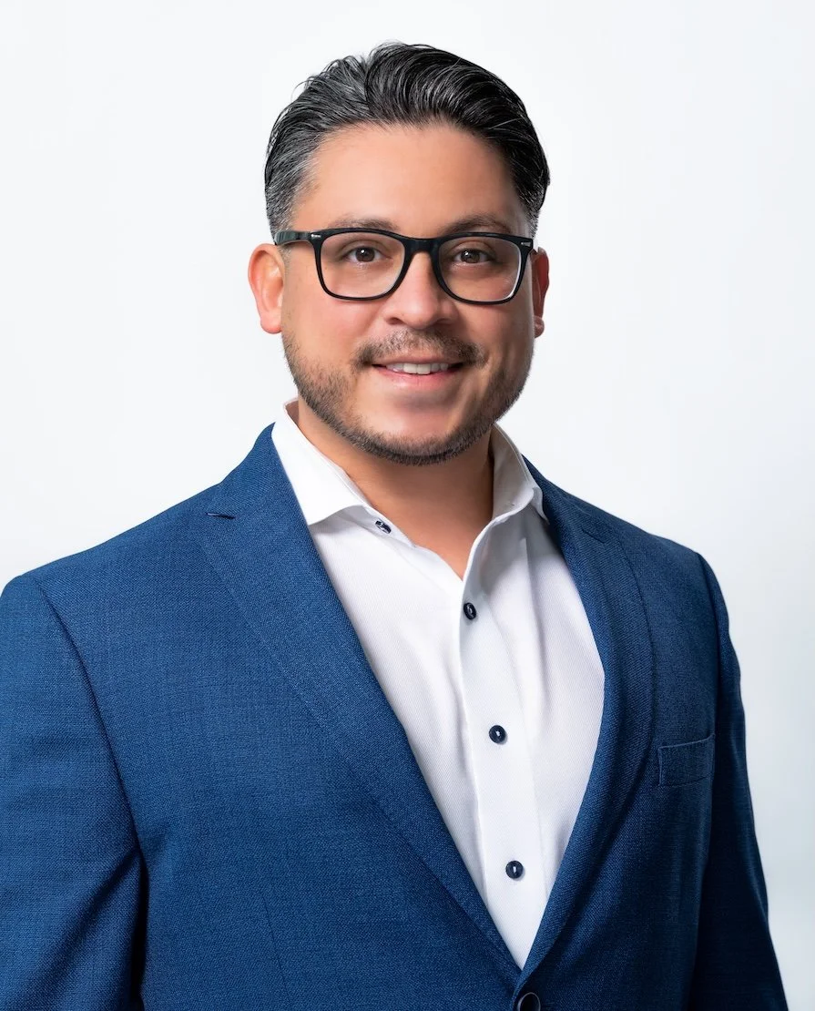 Professional Corporate Headshot by Saint Paul Photographer Jay Cupcake. Headshot of a man with dark hair, glasses, and light skin, dressed in a blue blazer and white shirt, smiling against a plain white background.