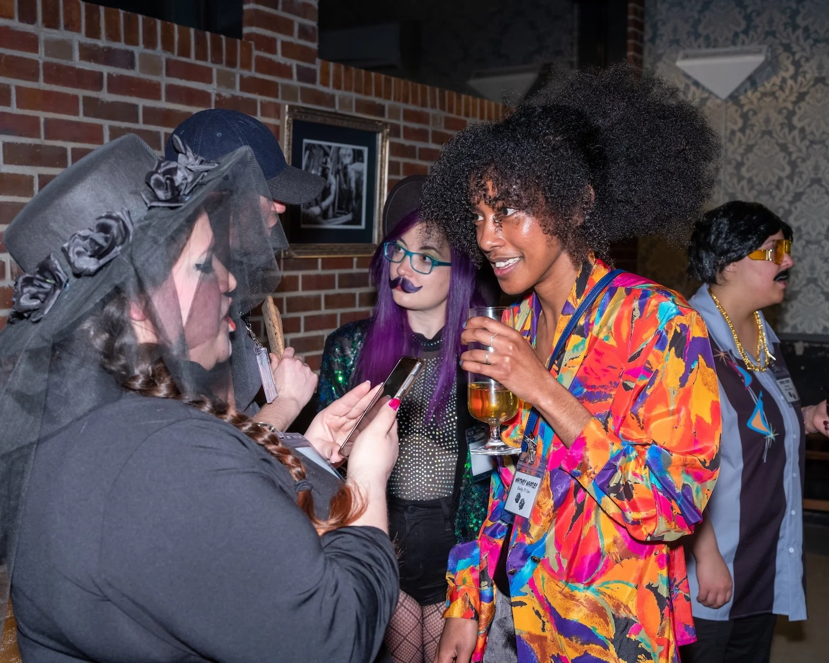 Professional Event Photography by Saint Paul Photographer Jay Cupcake. Group of women in colorful costumes socializing at a party, with brick wall and framed photo in background.