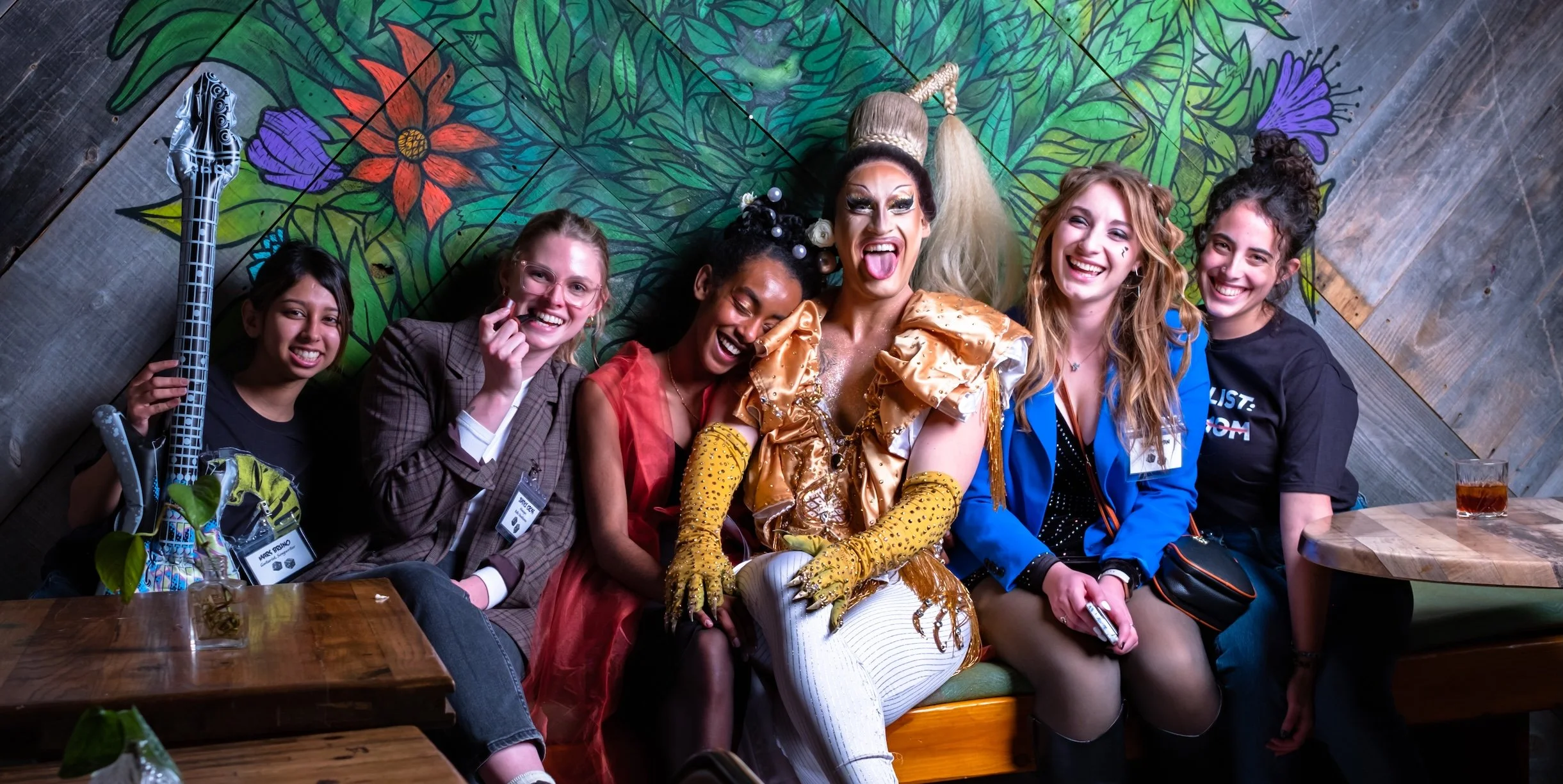 Professional Event Photography by Minneapolis Photographer Jay Cupcake. Group of six women sitting closely together in a restaurant, smiling, laughing, and making funny faces, with a colorful, floral mural wall behind them and tables with drinks in f