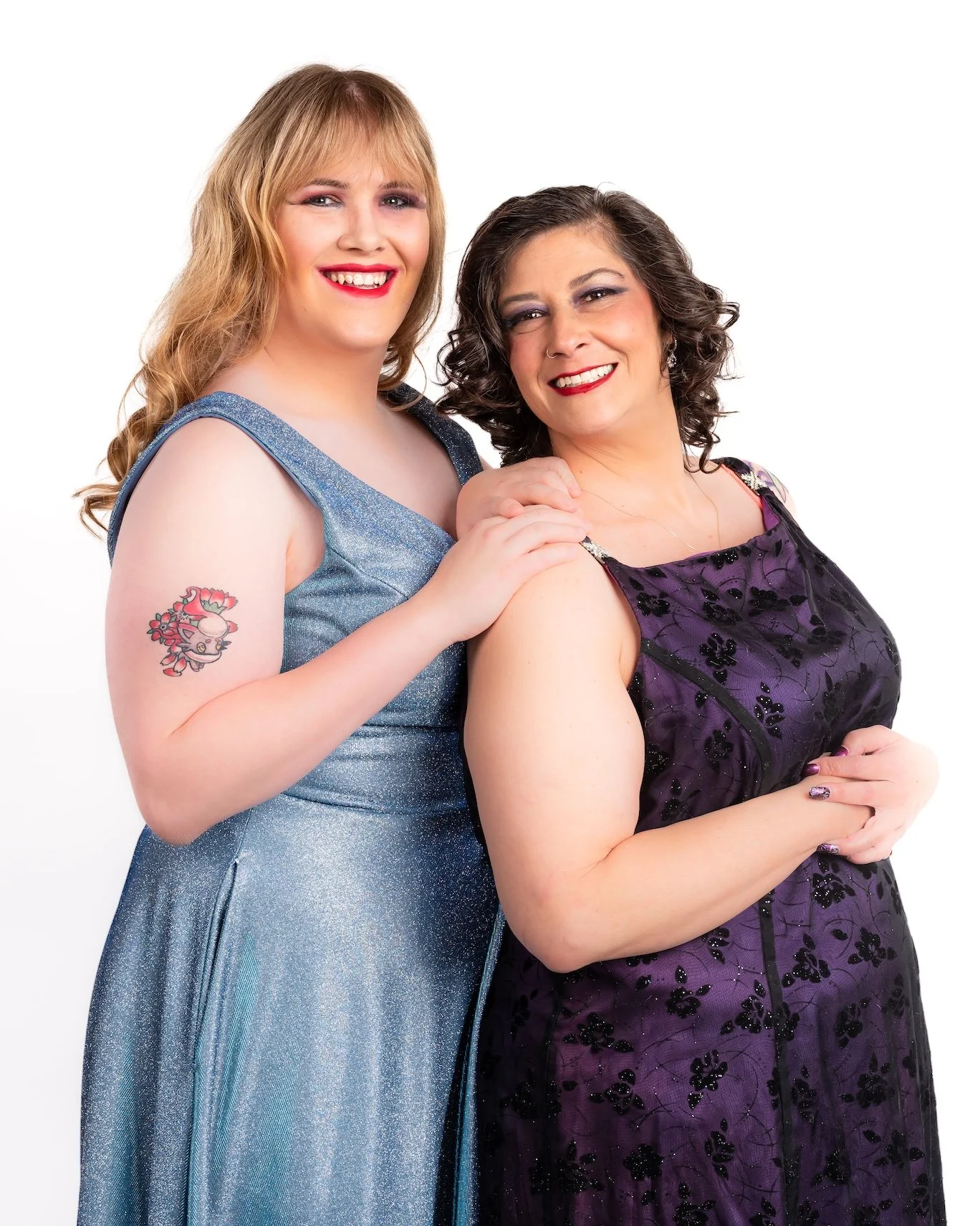 Professional Portrait Photography by Twin Cities Photographer Jay Cupcake. Two women smiling, one with blonde hair in a blue dress and the other with dark hair in a purple dress, posing together against a white background.