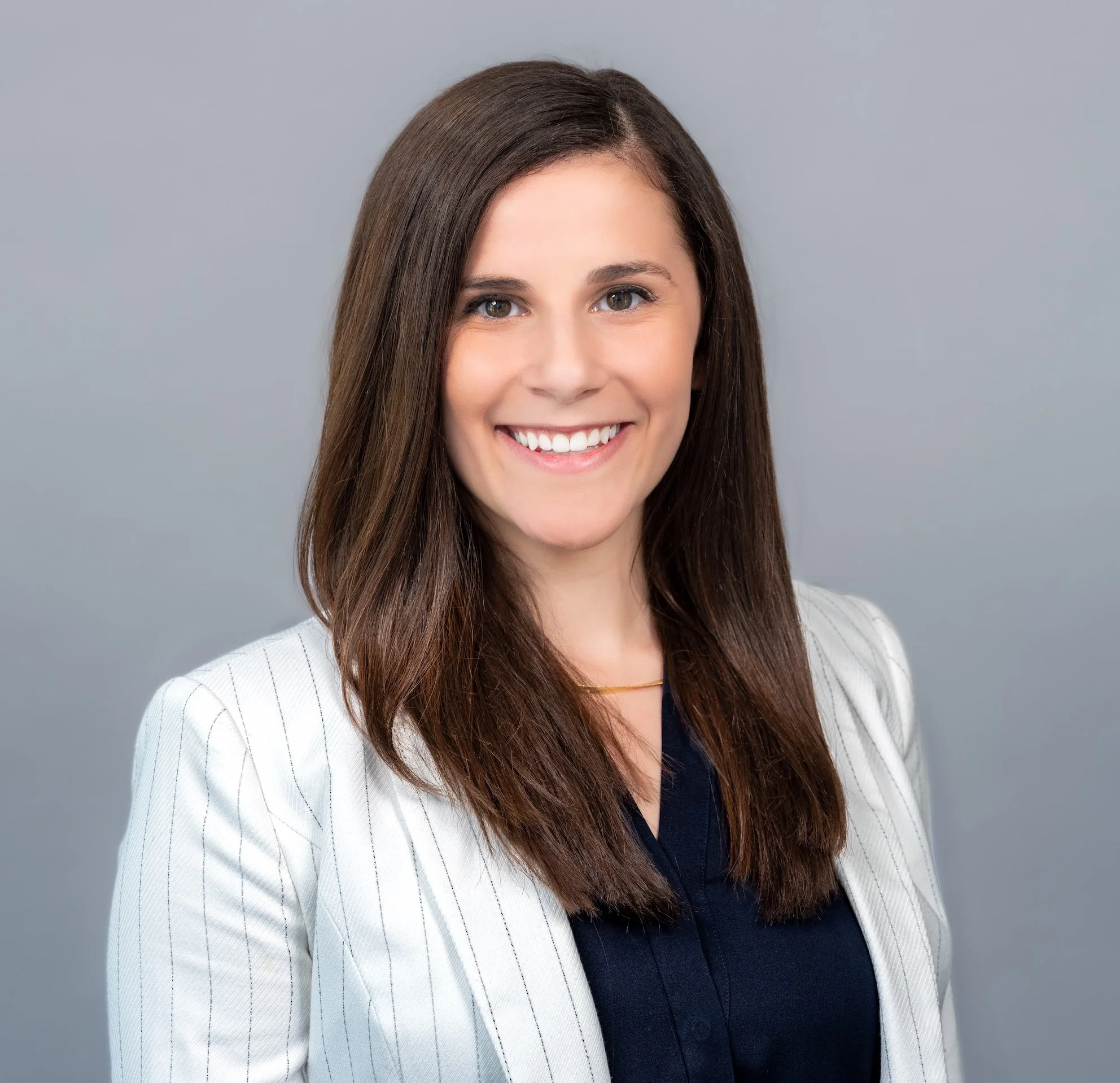 Professional Corporate Headshot by Minneapolis Photographer Jay Cupcake. A woman with long brown hair and blue eyes smiling at the camera, wearing a white pinstripe blazer over a dark top, against a plain gray background.
