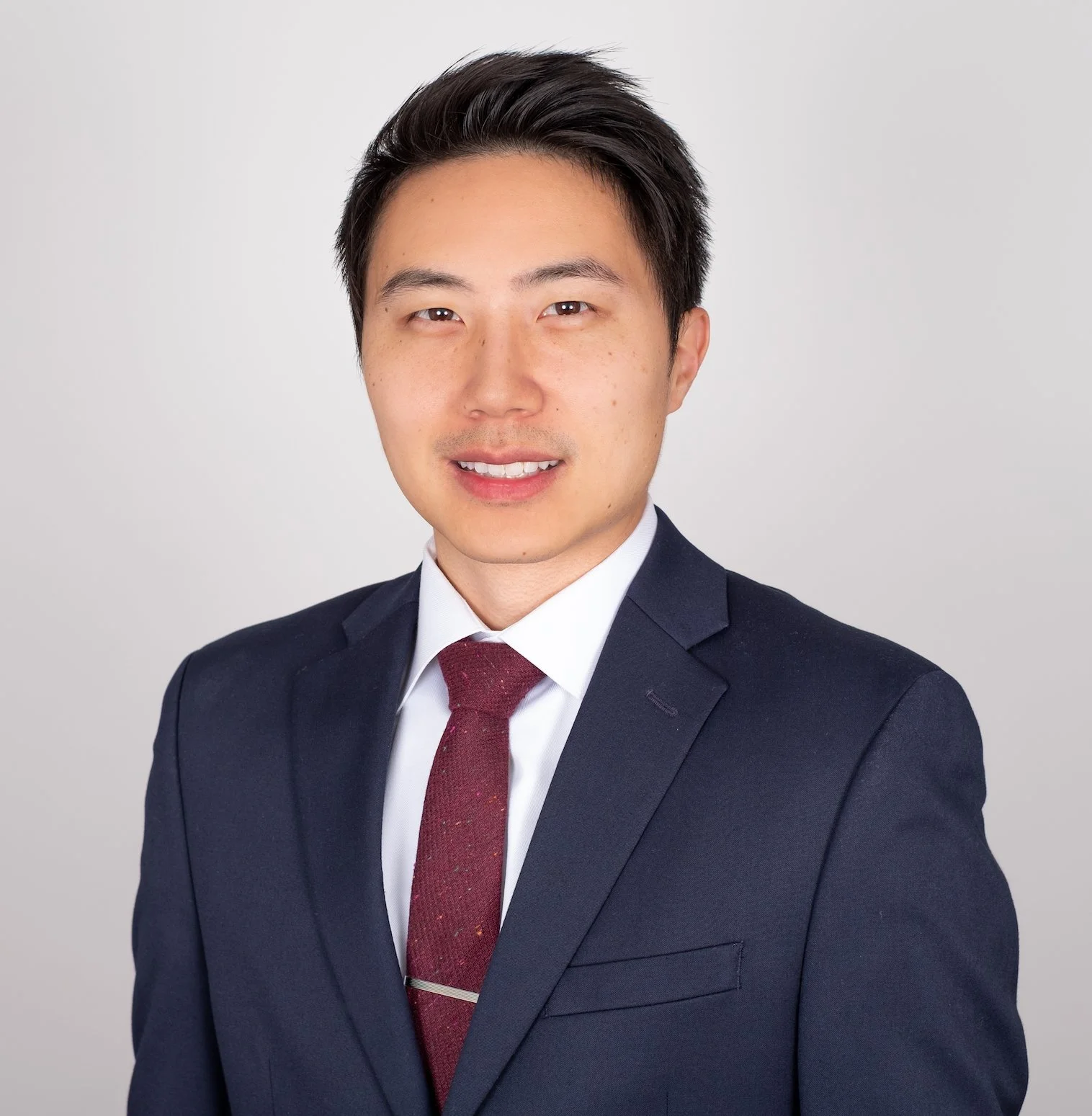 Professional Corporate Headshot by Twin Cities Photographer Jay Cupcake. Professional headshot of a young Asian man in a navy blue suit and maroon tie, smiling against a plain white background.