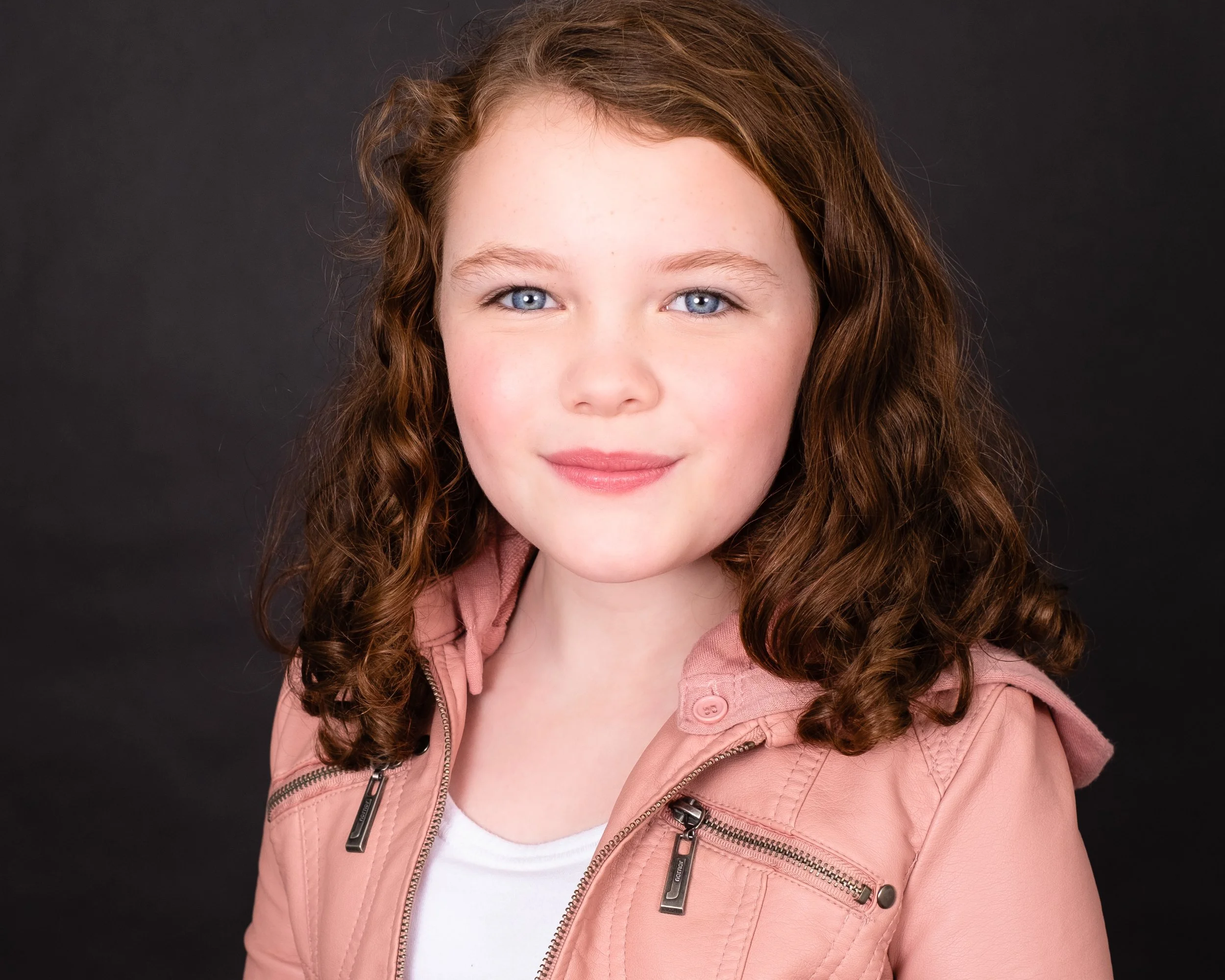 Professional headshot by St. Paul photographer Jay Cupcake. A young girl with curly red hair and blue eyes, wearing a light pink jacket and a white shirt, smiling slightly against a dark background.
