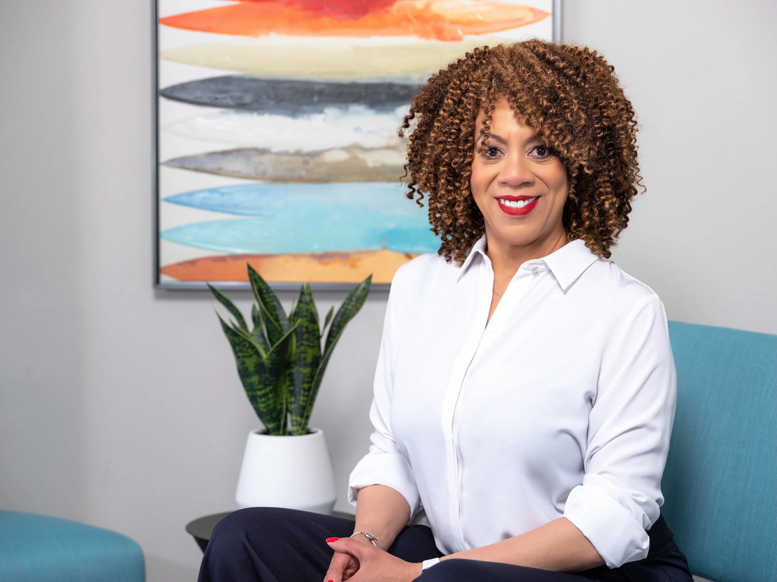 Professional Portrait Photography by Minneapolis Photographer Jay Cupcake. A woman with curly hair, wearing a white blouse, sitting on a blue chair in an office with a colorful abstract painting and a potted plant in the background.