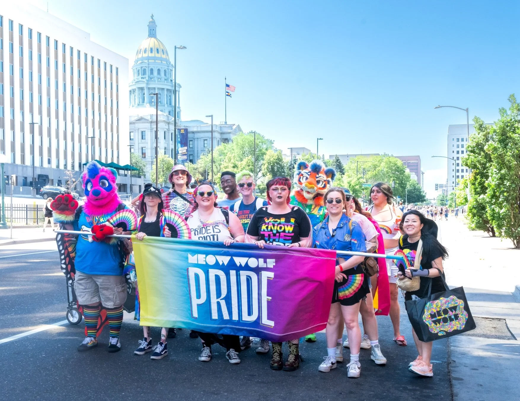 Professional Event Photography by Minneapolis Photographer Jay Cupcake. Group of people participating in a Pride parade, holding a rainbow-colored banner that reads 'Meow Wolf Pride', with some dressed in colorful costumes and accessories, walking on