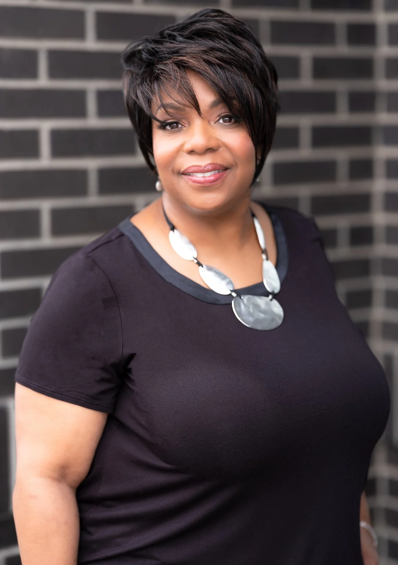 Professional Corporate Headshot by Twin Cities Photographer Jay Cupcake. Portrait of a woman with short, dark hair and a black top, standing in front of a brick wall.