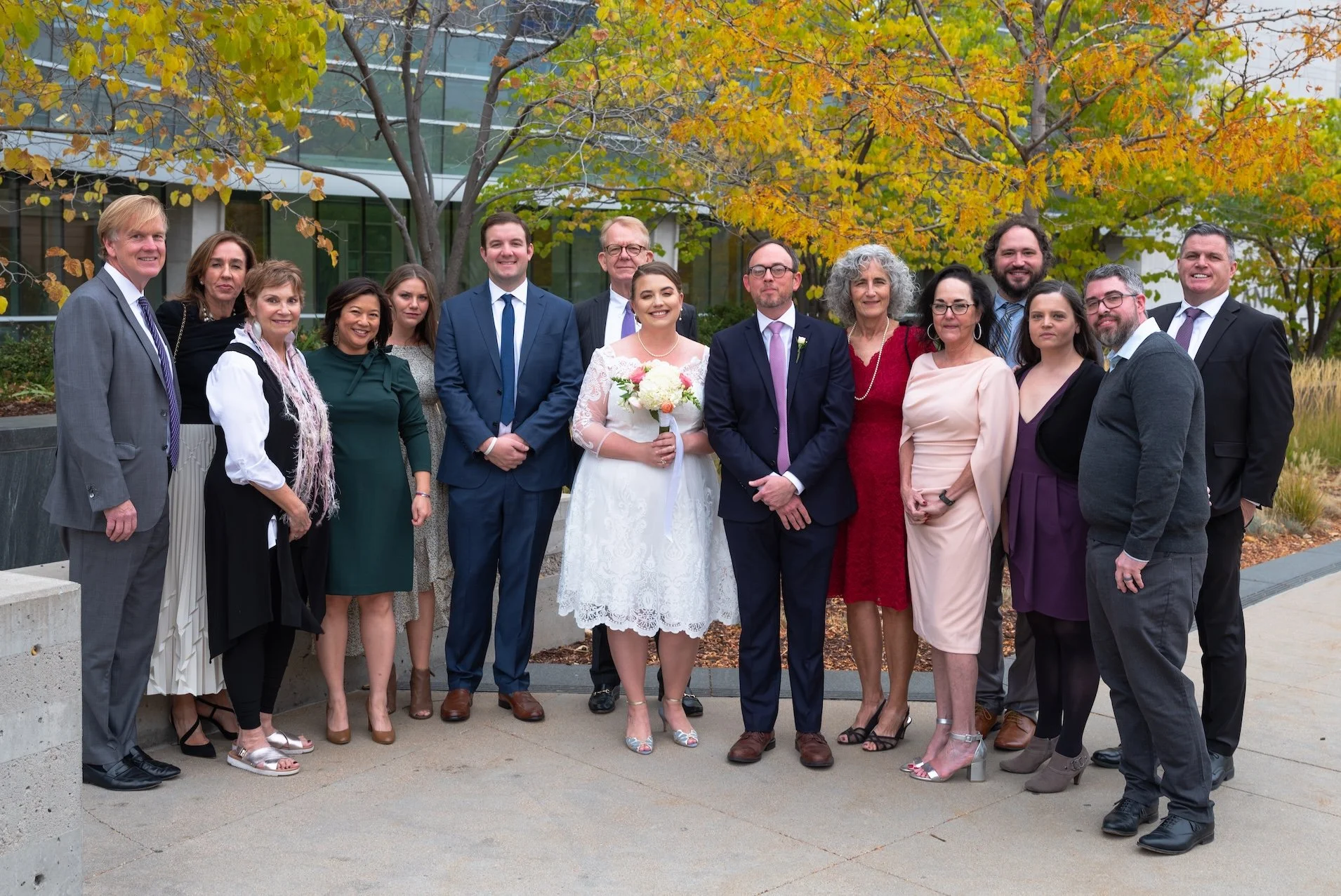 Professional Event Photography by Saint Paul Photographer Jay Cupcake. Group of people dressed in formal attire at a wedding outdoors, with trees and a modern building in the background.