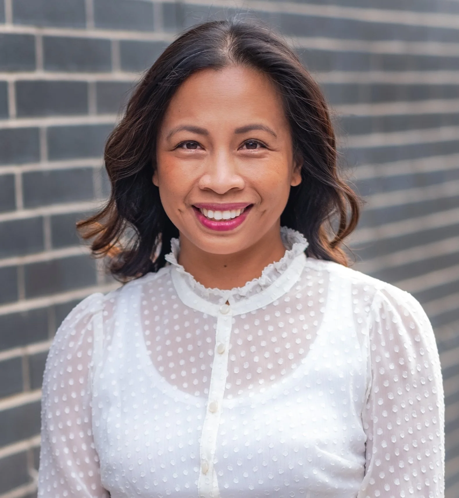 Professional Corporate Headshot by Saint Paul Photographer Jay Cupcake. Smiling woman with shoulder-length dark hair wearing a white polka dot blouse standing in front of a brick wall.