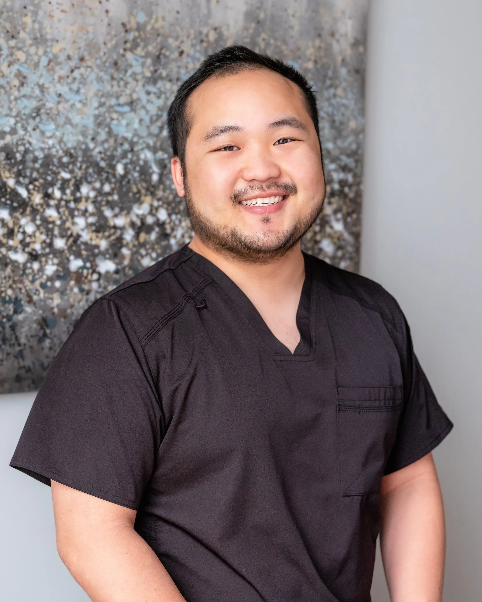 Professional Corporate Headshot by Minneapolis Photographer Jay Cupcake. A man wearing a black medical scrub top, smiling, with a textured abstract art piece behind him.