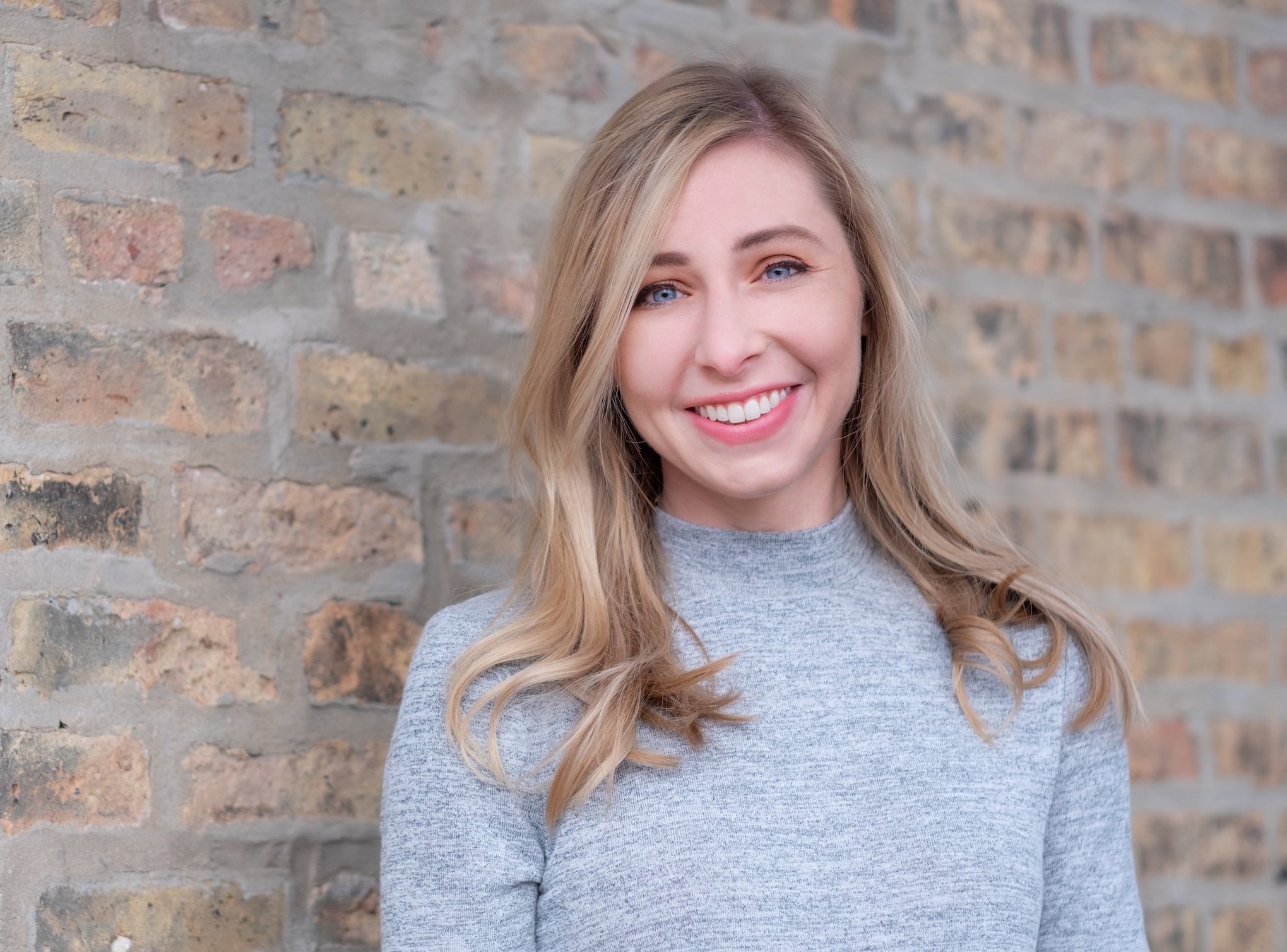 Professional Corporate Headshot by Twin Cities Photographer Jay Cupcake. Young woman with blonde hair and blue eyes smiling, wearing a gray sweater, standing in front of a brick wall.