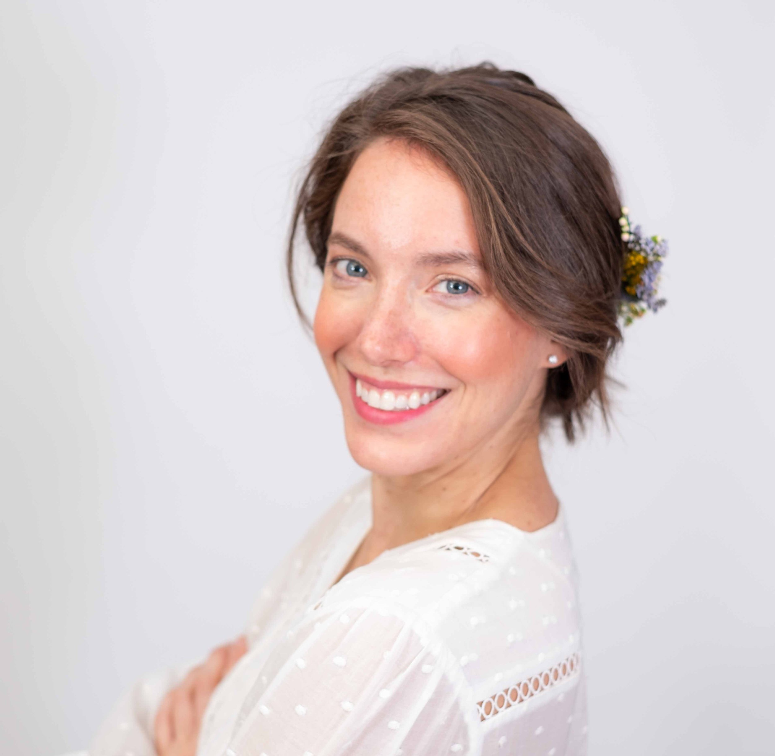 Professional Corporate Headshot by Twin Cities Photographer Jay Cupcake. A woman with short brown hair smiling with a flower tucked behind her ear, wearing a white blouse, in front of a plain background.