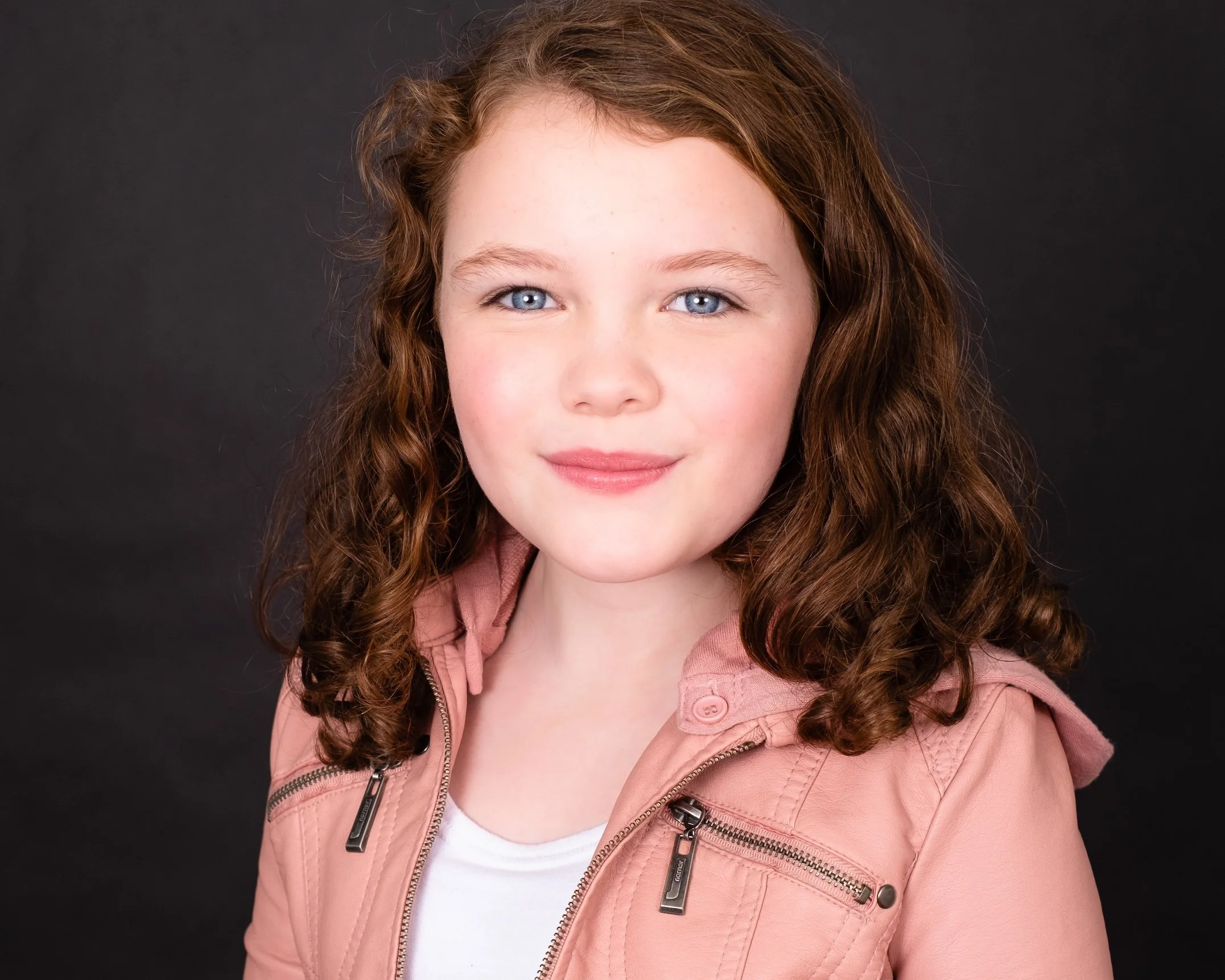 Professional Actor Headshot by Twin Cities Photographer Jay Cupcake. Portrait of a young girl with curly red hair and blue eyes, wearing a pink jacket and white shirt, smiling slightly against a black background.