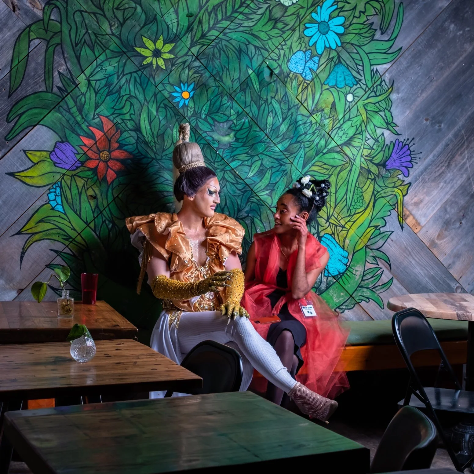 Professional Event Photography by Saint Paul Photographer Jay Cupcake. Two women sitting on a bench in front of a colorful floral mural. The woman on the left wears a gold and white outfit with gloves, and has a tall, elaborate hairstyle. The woman o