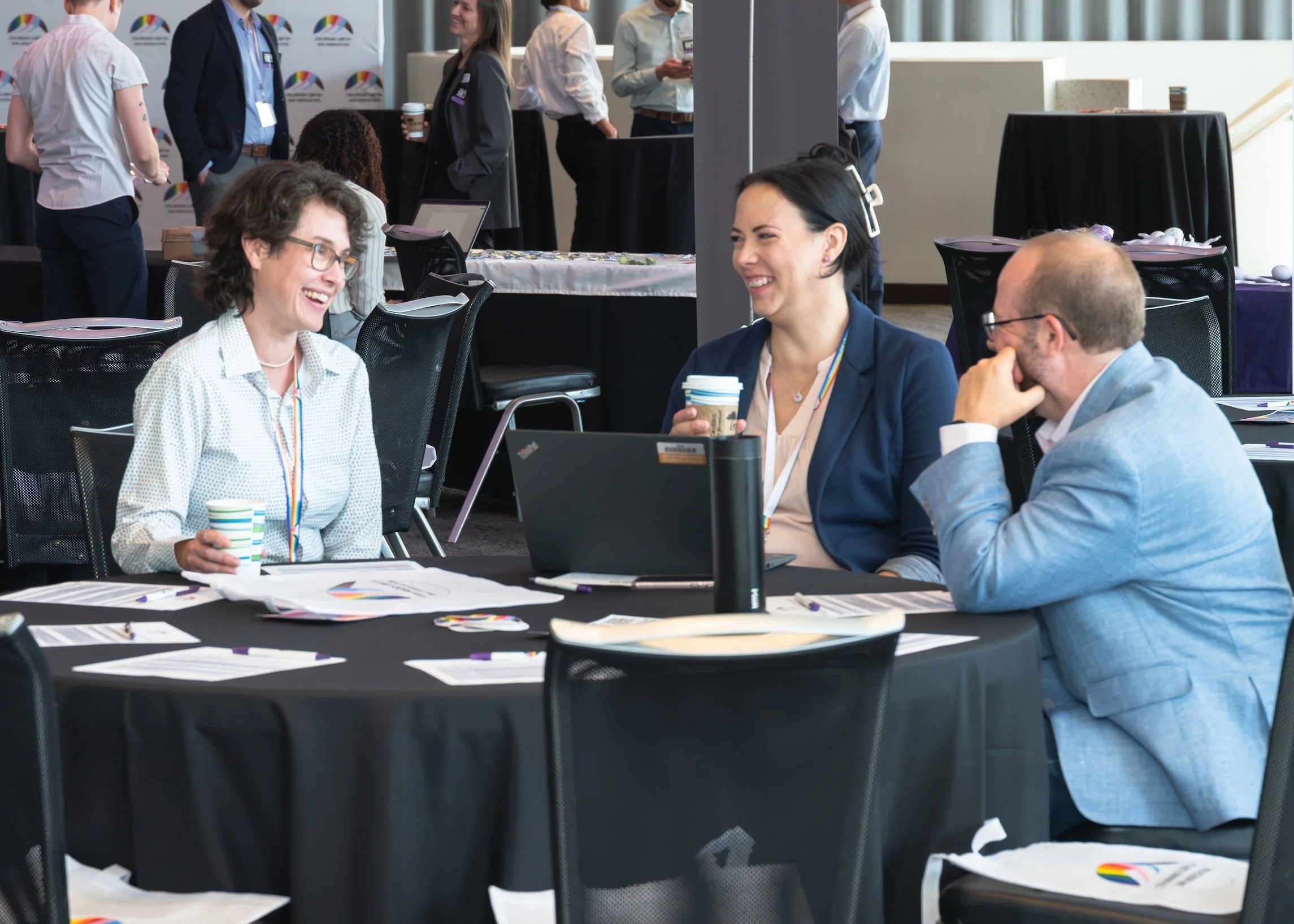 Professional Event Photography by Minneapolis Photographer Jay Cupcake. Three professionals sitting at a conference table, engaged in conversation and smiling. The woman on the left has curly hair and glasses, holding a paper cup. The woman in the mi