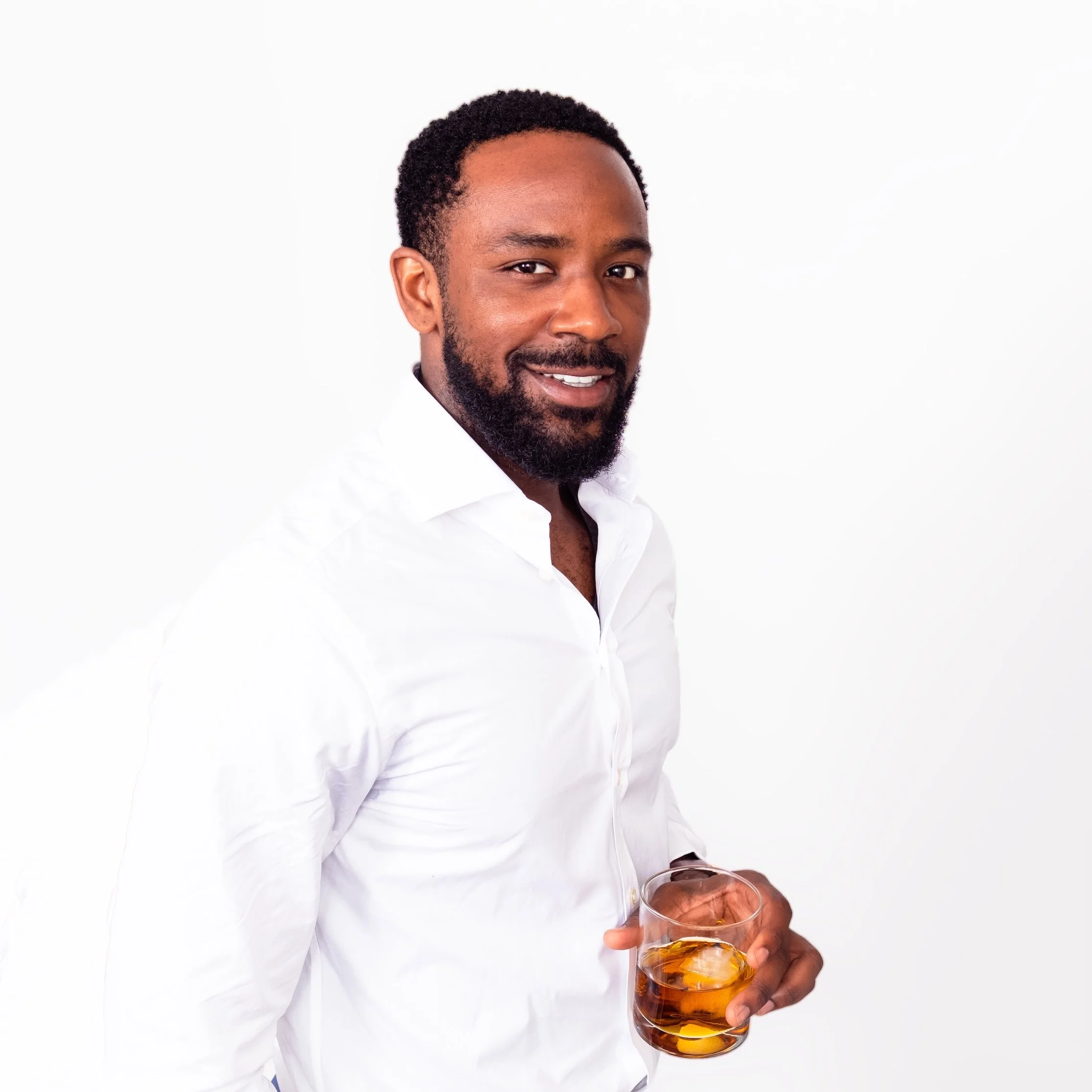 Professional Portrait Photography by Minneapolis Photographer Jay Cupcake. Man with dark curly hair and beard wearing a white shirt, holding a glass of whiskey, smiling, standing against a white background.