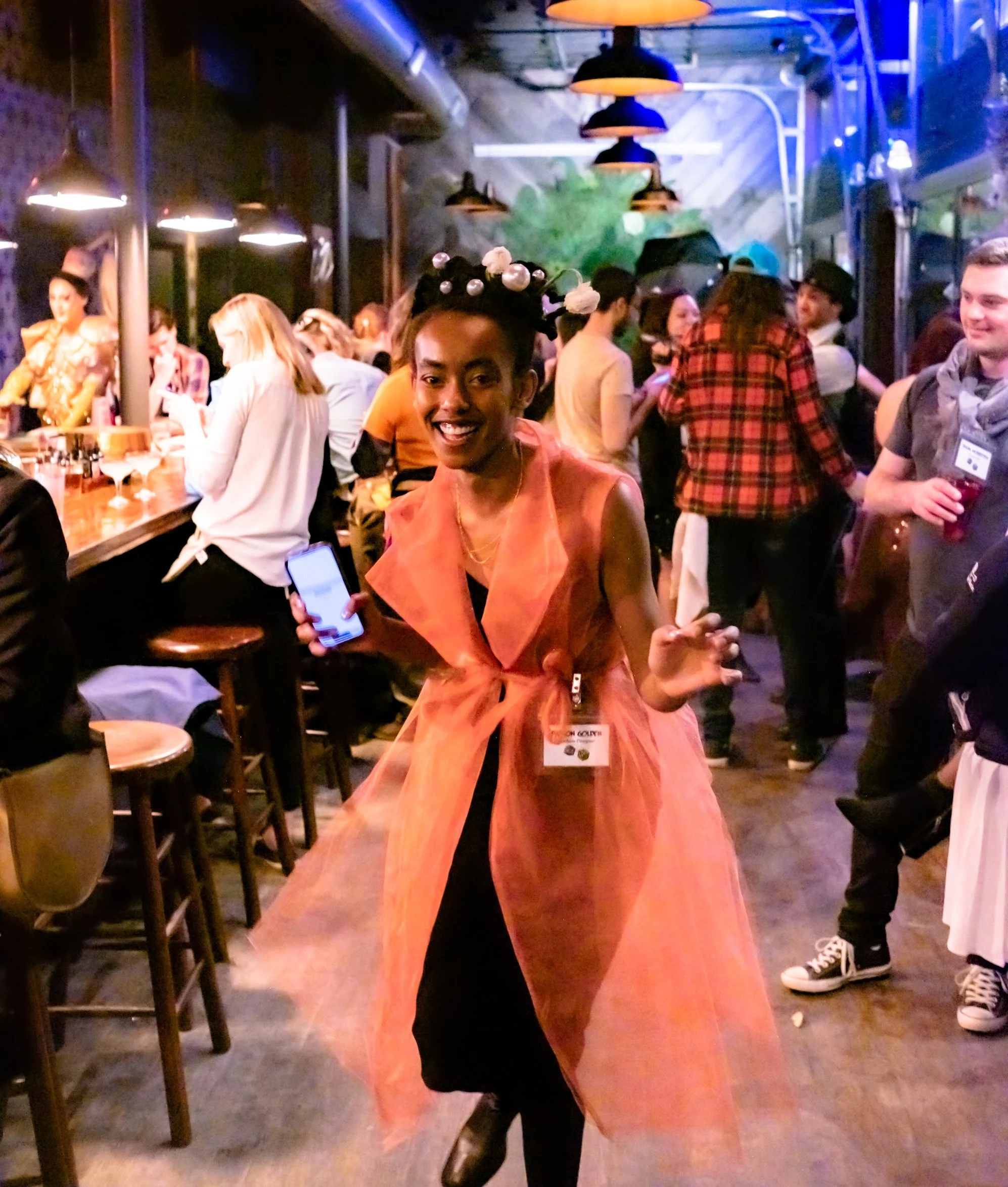 Professional Event Photography by Saint Paul Photographer Jay Cupcake. A woman with a joyful expression wearing an orange sleeveless dress, black tights, and a unique headpiece with pearls, standing in a lively bar or restaurant setting among people 