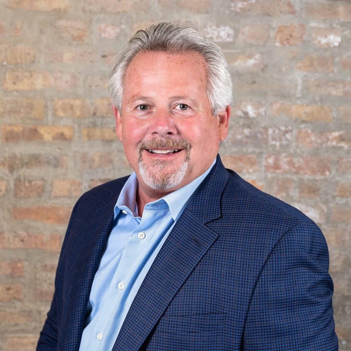 Professional Corporate Headshot by Saint Paul Photographer Jay Cupcake. A smiling middle-aged man with gray hair and a beard, wearing a blue suit jacket and light blue shirt, standing in front of a brick wall.