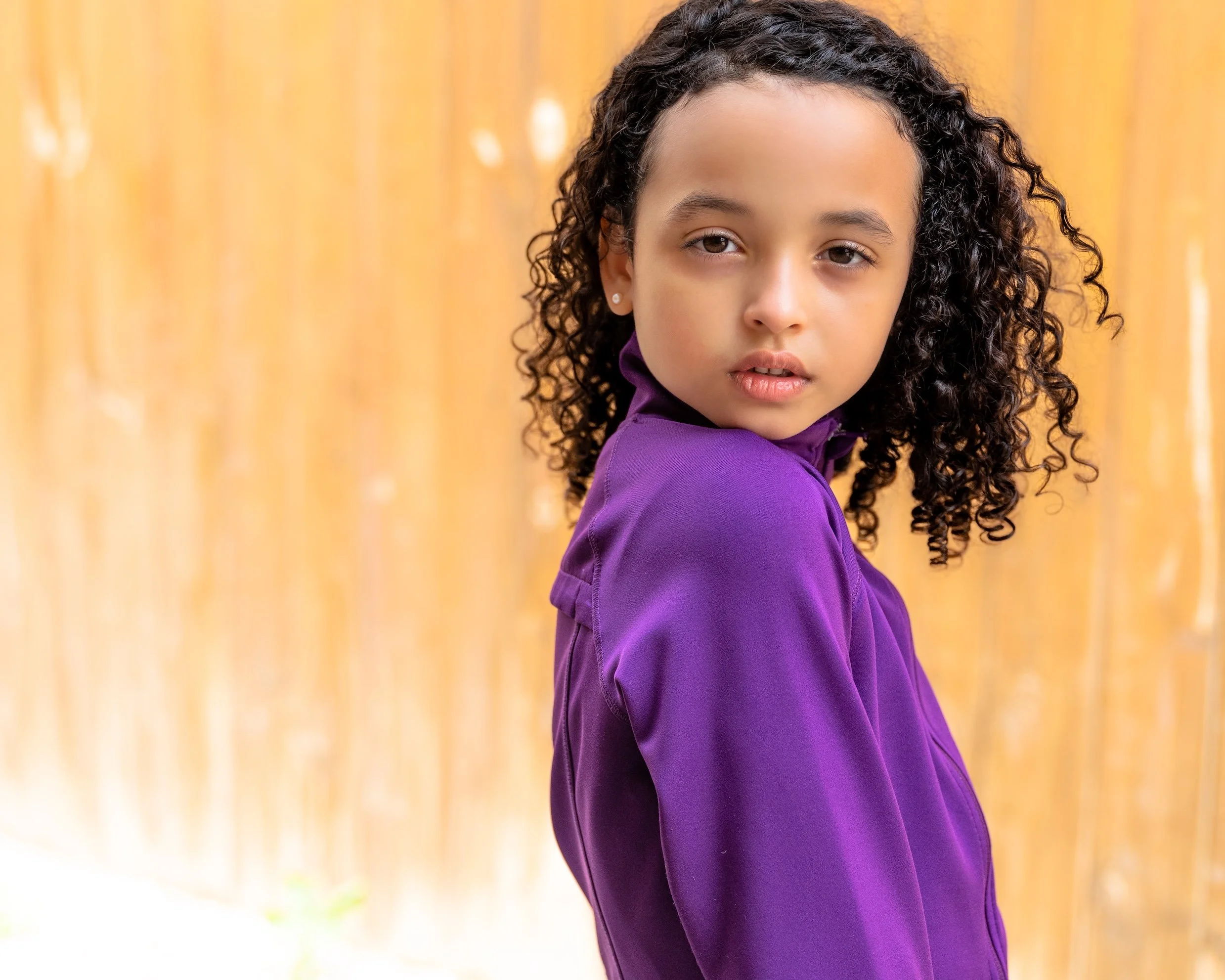 Professional Actor Headshot by Minneapolis Photographer Jay Cupcake. Young girl with curly hair wearing a purple jacket, standing outdoors with a wooden fence in the background.