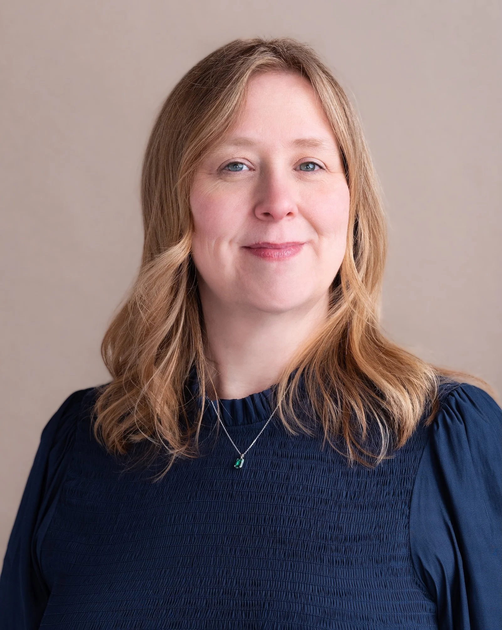 Professional Corporate Headshot by Minneapolis Photographer Jay Cupcake. Close-up portrait of a woman with shoulder-length reddish-blonde hair and blue eyes, wearing a navy blue top and a silver necklace with green gemstone pendant, against a neutral