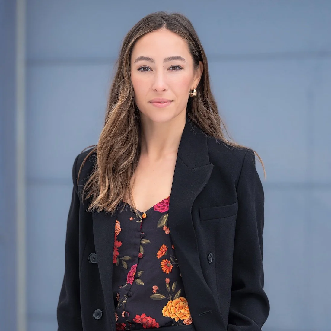 Professional Portrait Photography by Saint Paul Photographer Jay Cupcake. A woman with long wavy brown hair, wearing a black blazer and a dark floral blouse, standing outdoors against a blue background.