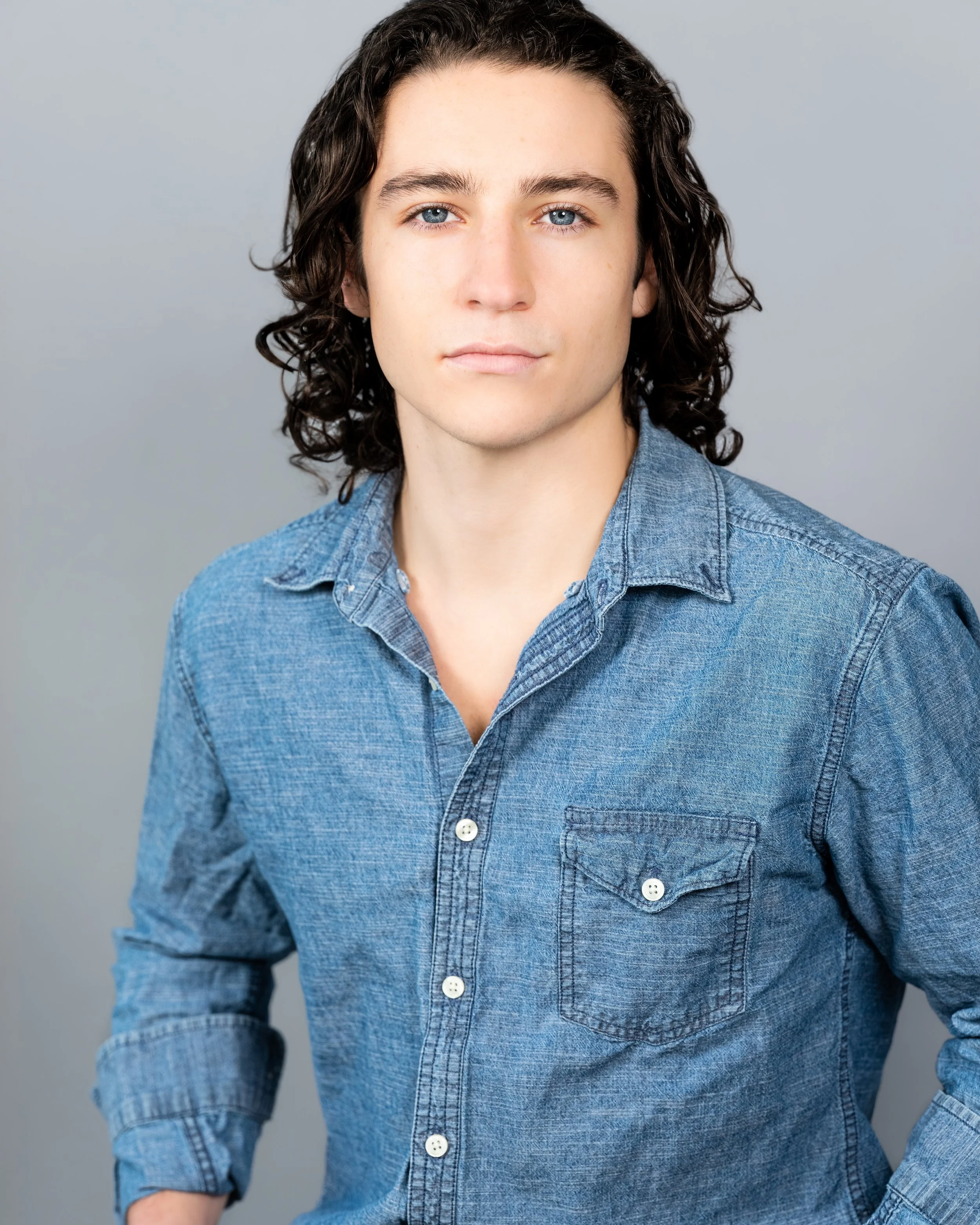 Professional headshot by St. Paul photographer Jay Cupcake. Portrait of a young man with shoulder-length curly dark hair, blue eyes, wearing a denim button-up shirt, standing against a plain gray background.