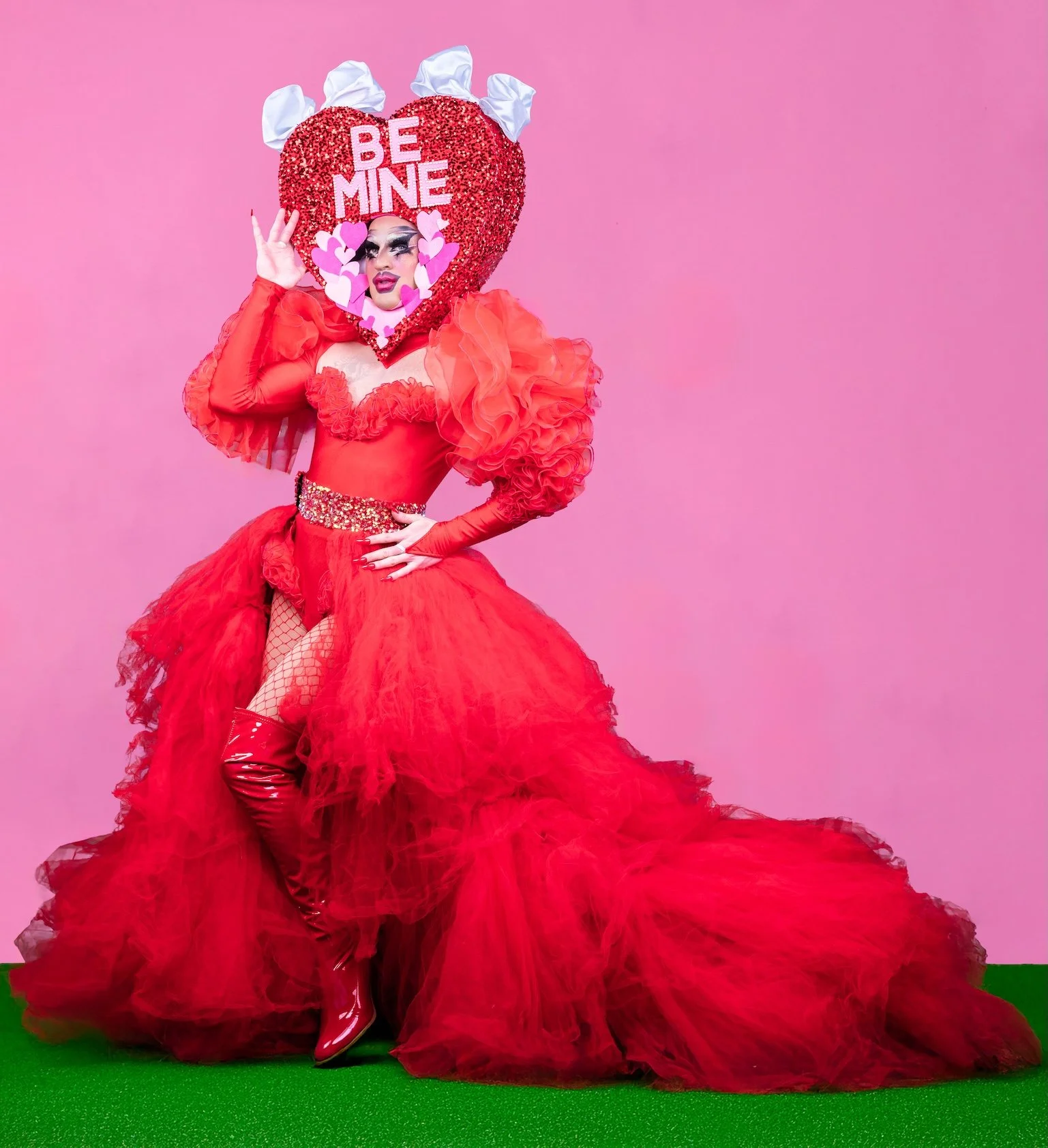 Professional Portrait Photography by Twin Cities Photographer Jay Cupcake. Person in a red dress with puffy sleeves, fishnet stockings, and thigh-high red patent boots, wearing a large heart-shaped headpiece with the words 'BE MINE' and pink hearts.