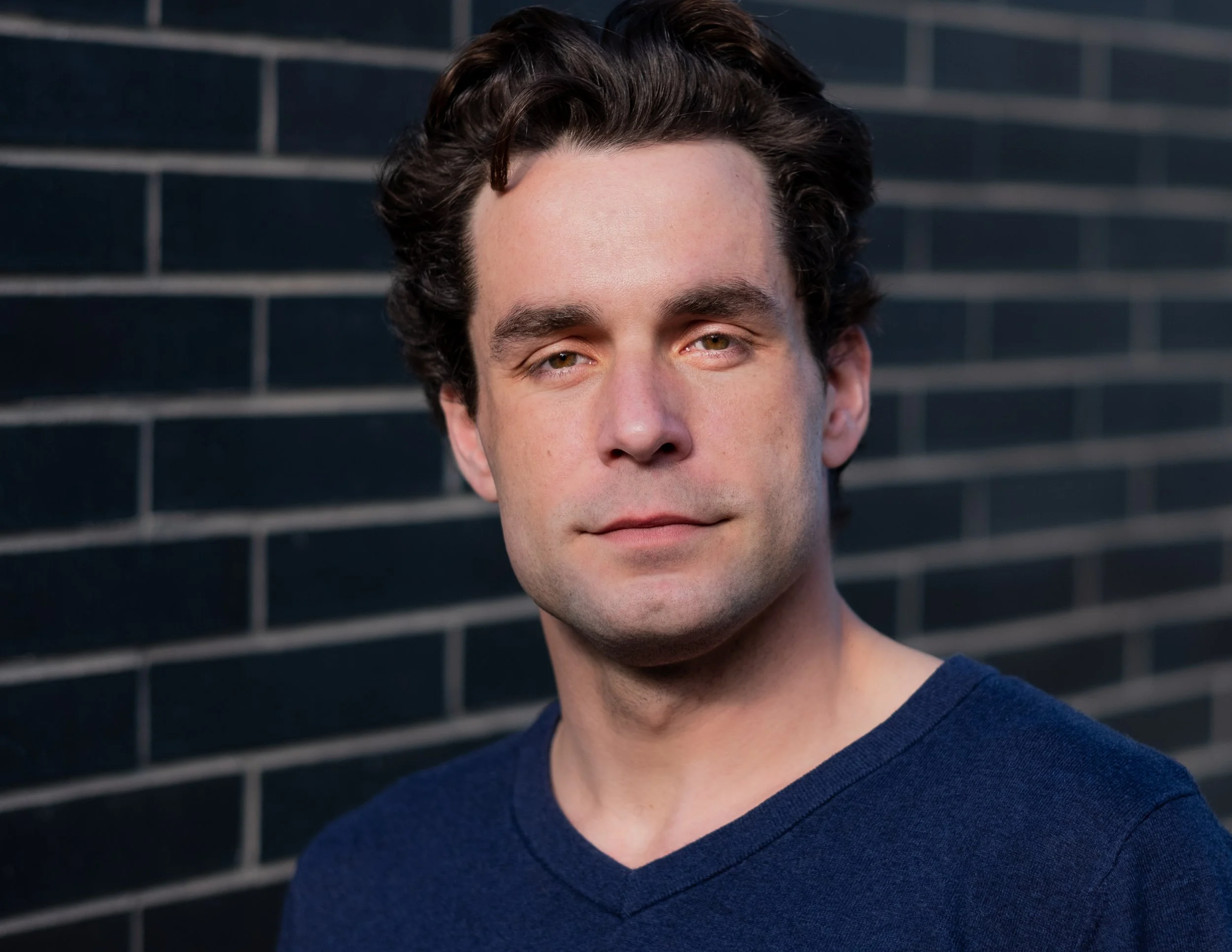 Professional headshot by St. Paul photographer Jay Cupcake. Close-up portrait of a man with dark curly hair, wearing a navy blue shirt, standing in front of a dark brick wall.