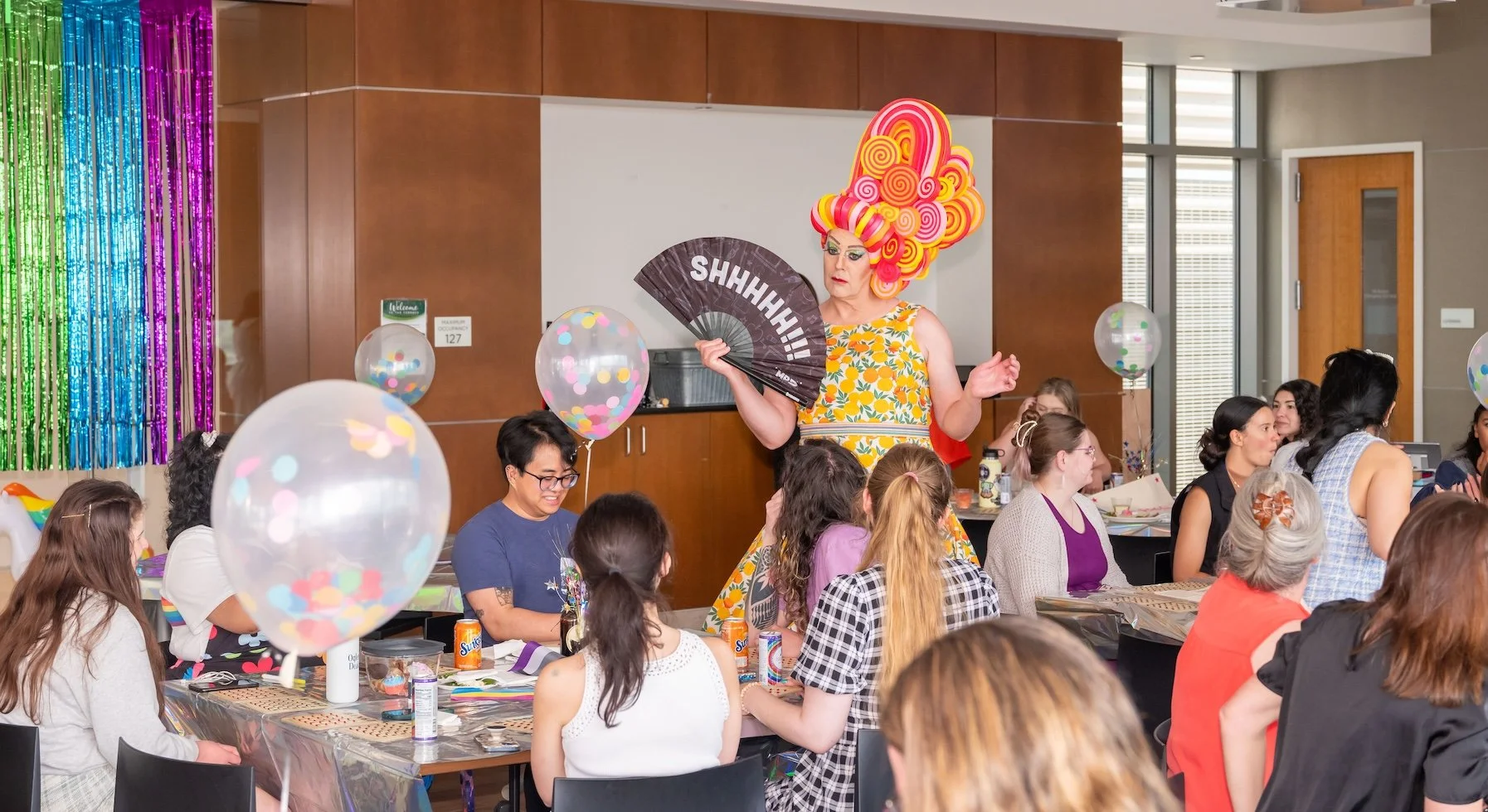 Professional Event Photography by Saint Paul Photographer Jay Cupcake. A woman with a large, colorful, swirly pink and yellow wig stands with a black fan that says "SHHH!!!" in a room decorated with rainbow streamers and balloons, while people sit at