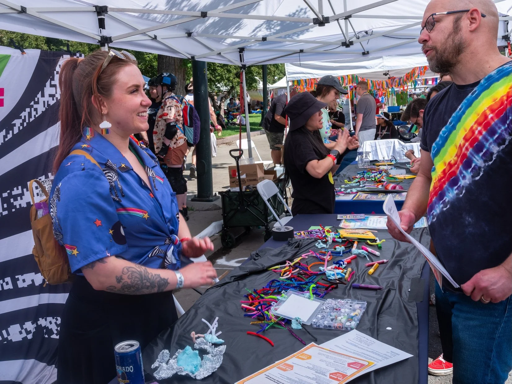 Professional Event Photography by Twin Cities Photographer Jay Cupcake. A woman with a rainbow-themed shirt and colorful earrings is smiling and talking to a man with glasses and a rainbow tie-dye shirt at a booth with various colorful craft supplies