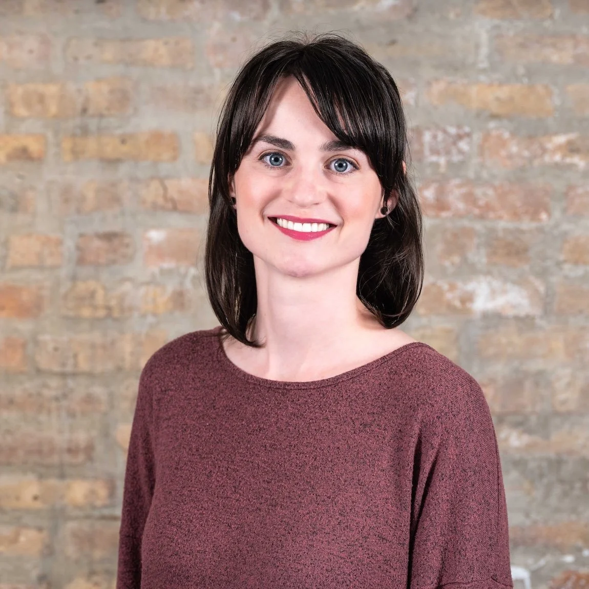 Professional Corporate Headshot by Minneapolis Photographer Jay Cupcake. A young woman with shoulder-length dark hair, blue eyes, and fair skin, smiling, wearing a maroon sweater, standing in front of a brick wall.