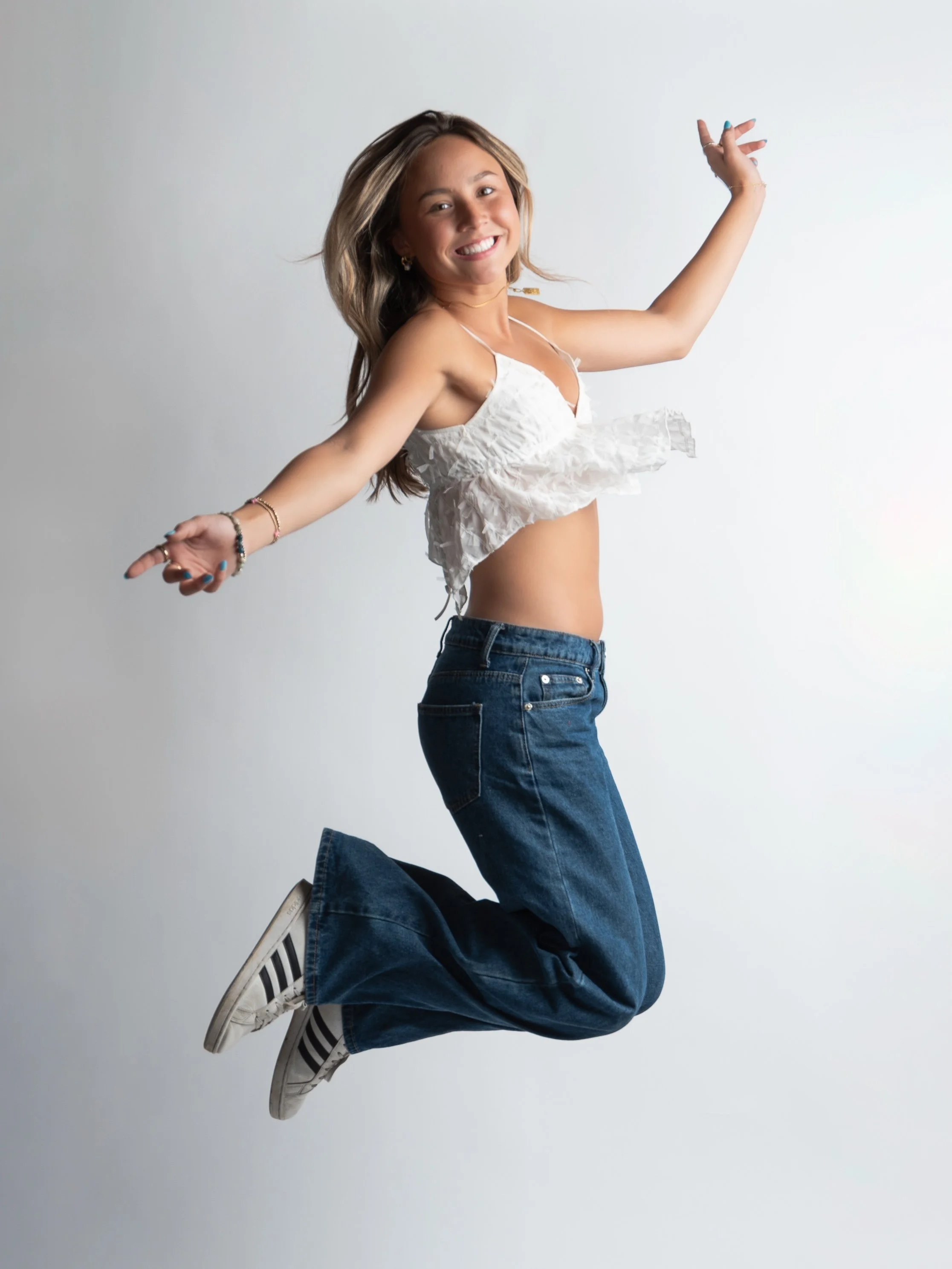 Professional Portrait Photography by Twin Cities Photographer Jay Cupcake. A young woman is jumping in the air with her arms outstretched and smiling. She is wearing a white ruffled crop top, blue jeans, and white sneakers with black stripes, against